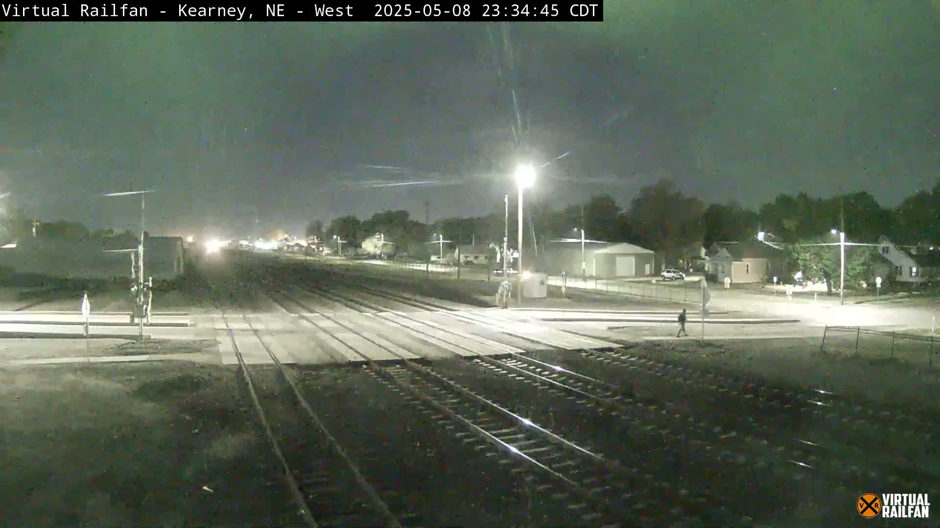 A nighttime view of railroad tracks intersecting a road, with a few buildings and trees visible in the background under a clear sky.