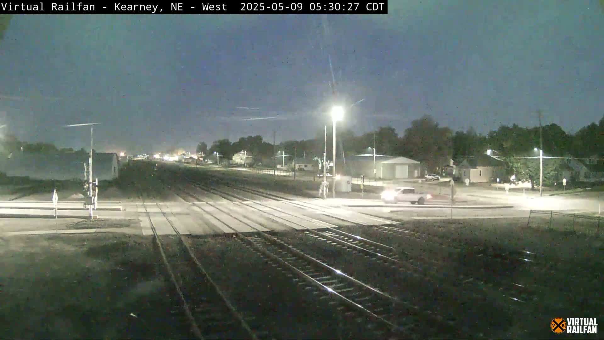 A nighttime view of a street crossing railroad tracks, with a single car visible on the road and trees and buildings in the background under a clear, dark sky.