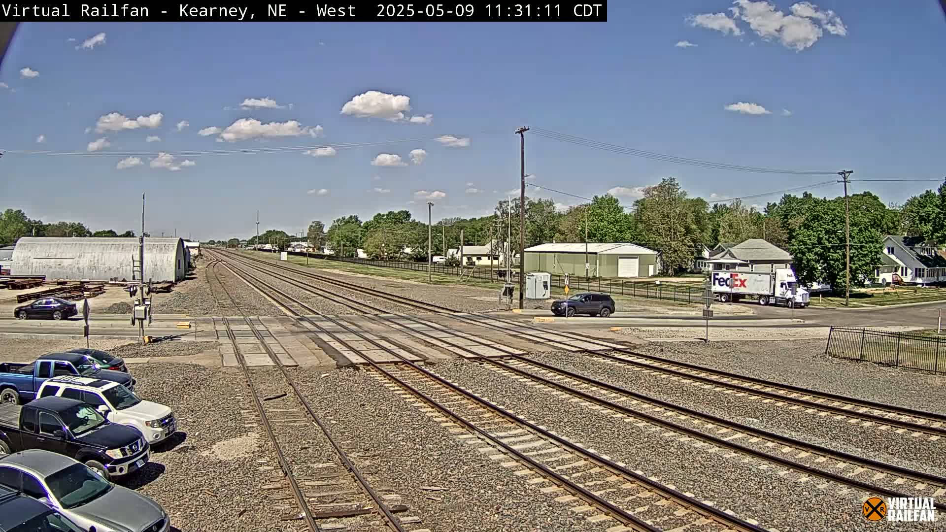 A sunny day reveals multiple sets of train tracks intersecting a road, with several vehicles parked and driving near a crossing, and buildings in the background.