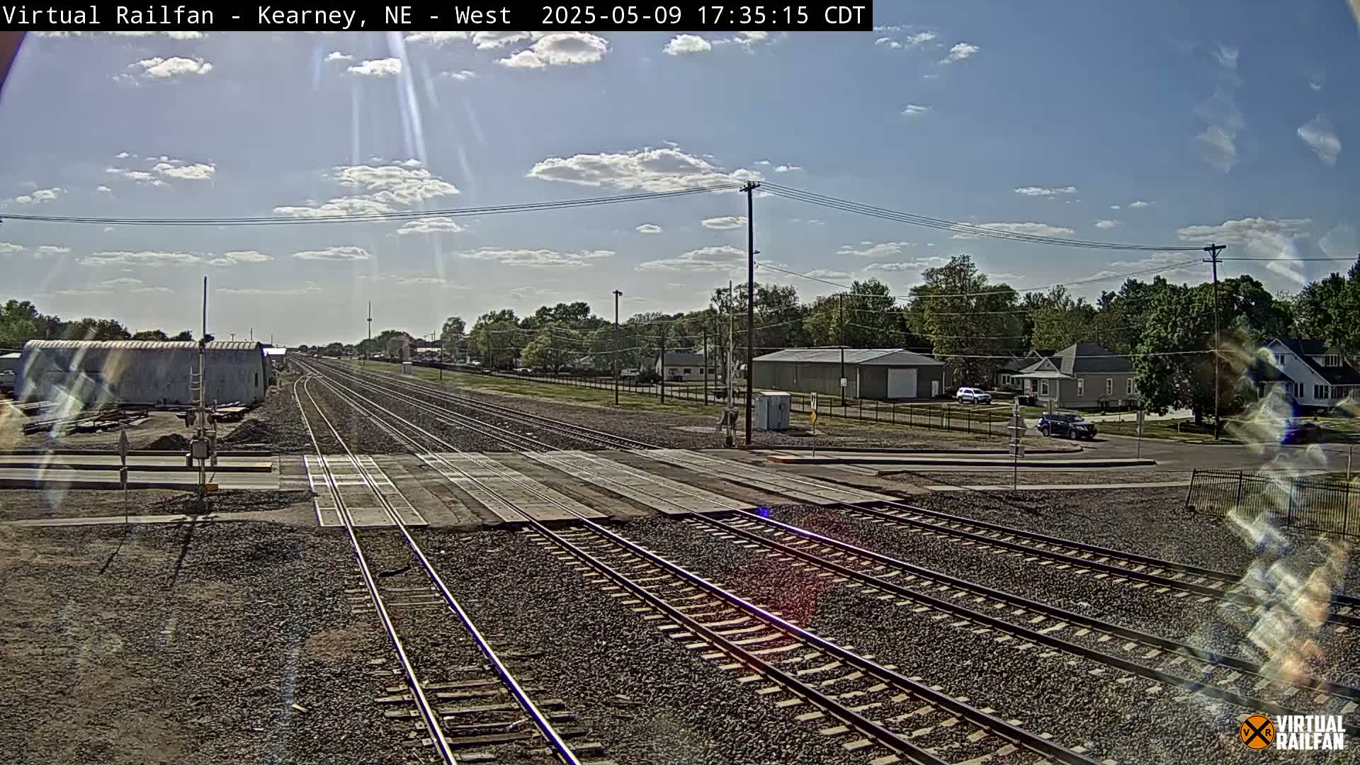 A sunny day with partly cloudy skies shows a railroad crossing in a residential area with several buildings and trees visible in the background.