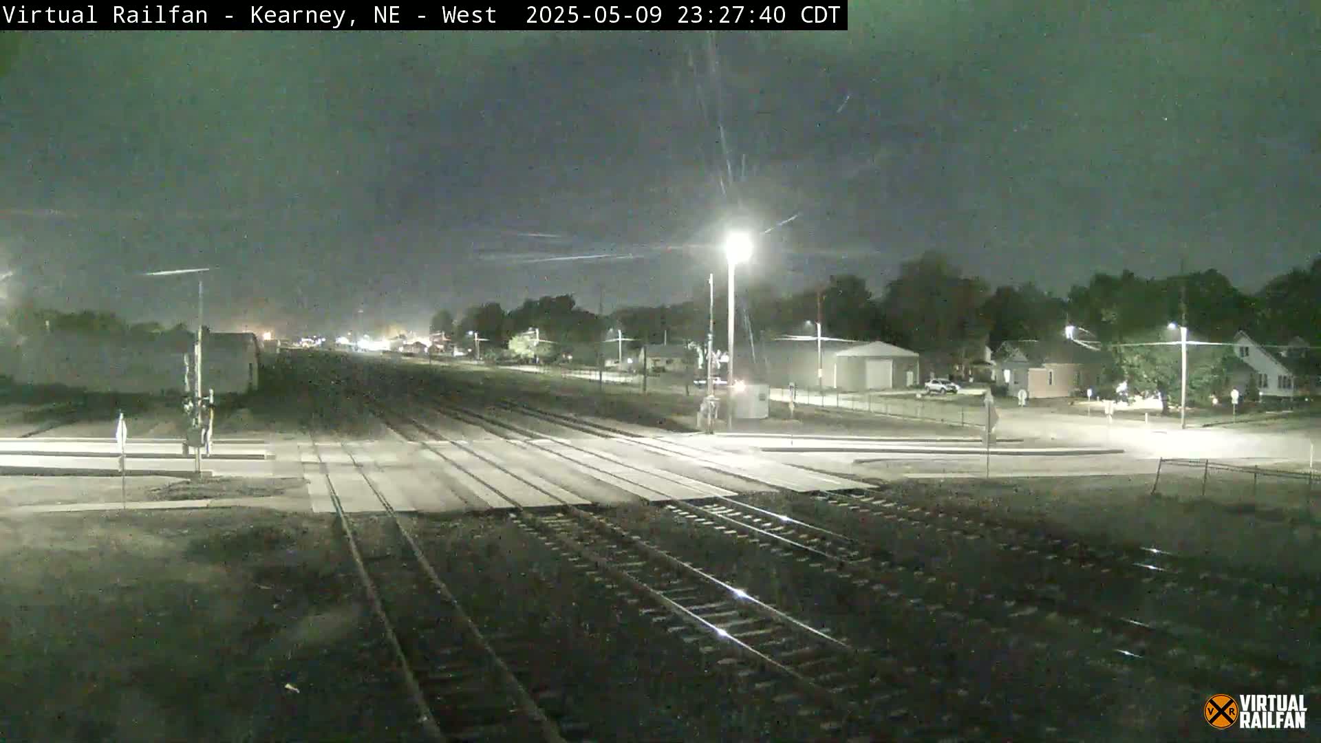 A nighttime view of railroad tracks intersecting a road, with residential buildings visible in the background under a dark, clear sky.