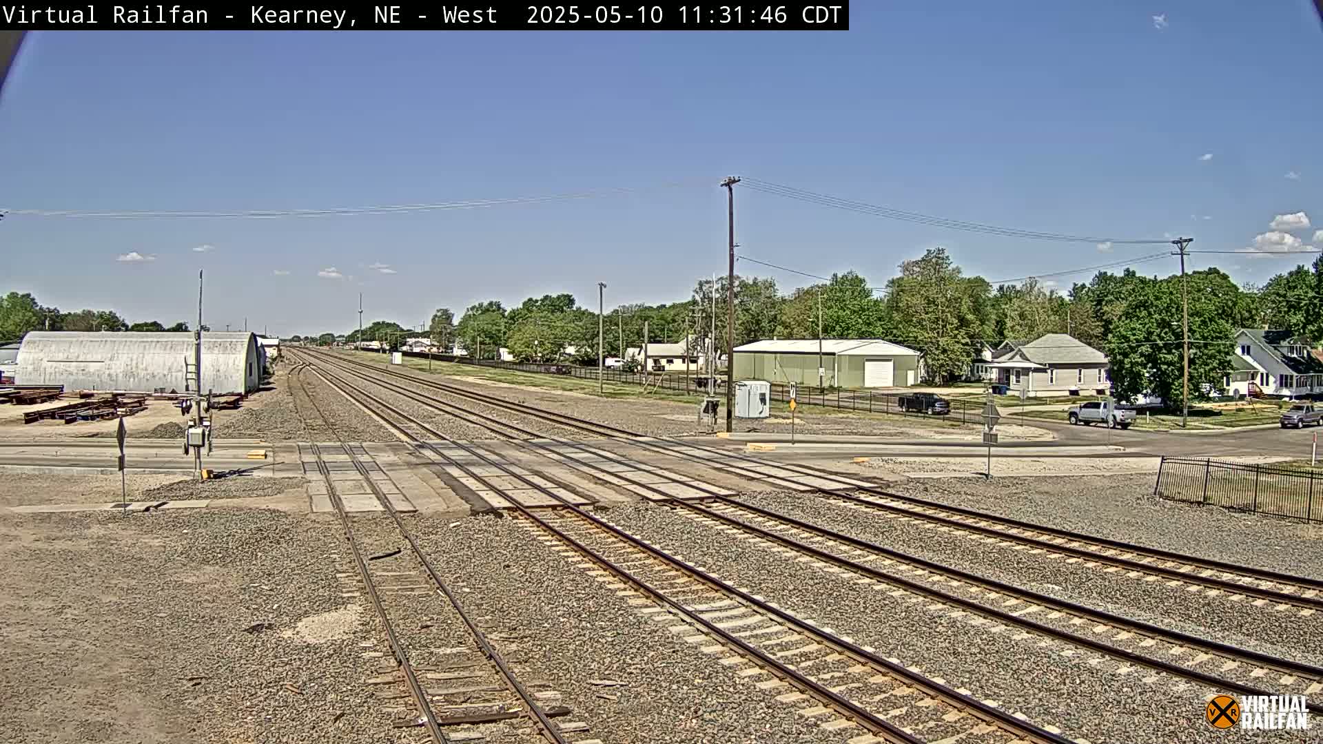 A railroad crossing in a small town on a sunny day, with several buildings and trees visible in the background.