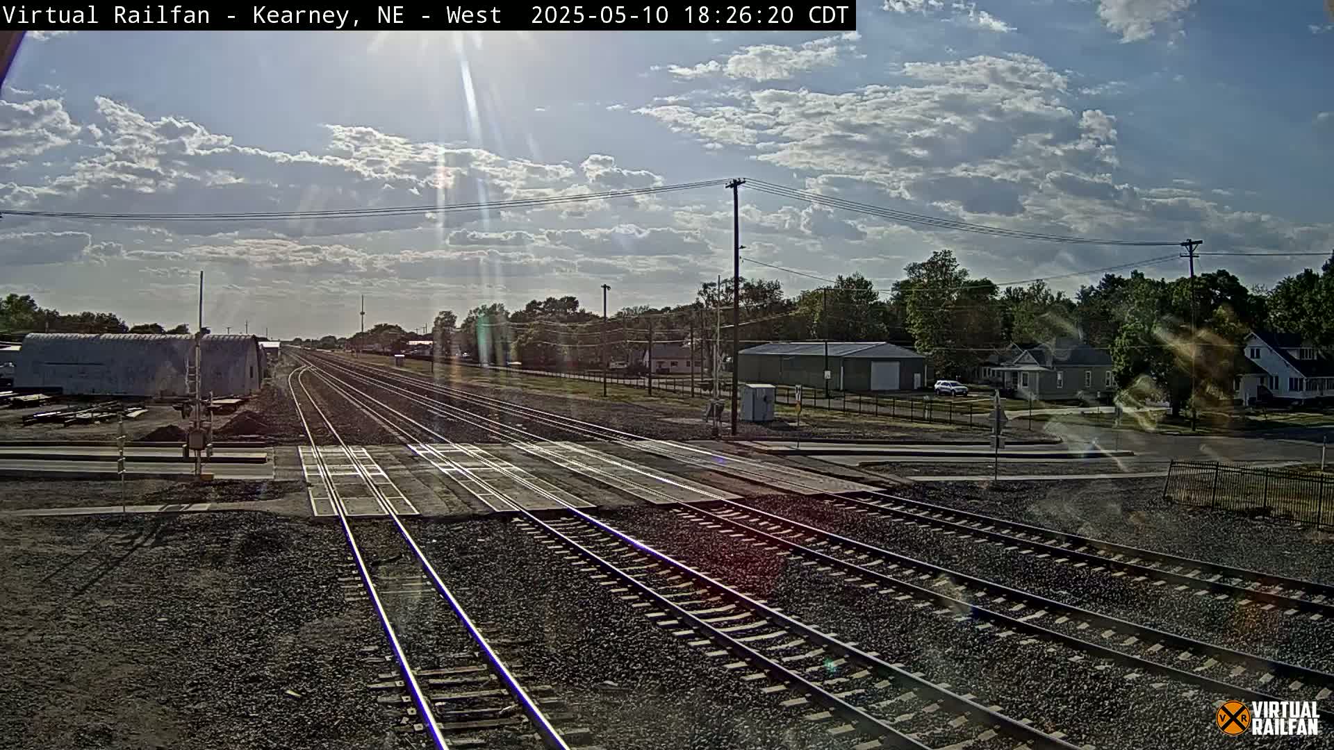 A sunny day with partly cloudy skies shows a view down several train tracks that intersect with a road, passing by residential buildings and industrial structures.