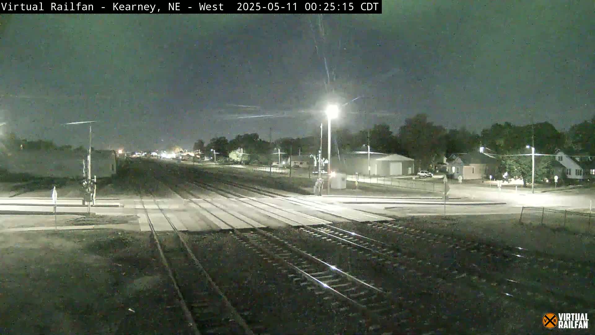 A nighttime view of railroad tracks intersecting a road, with residential buildings and streetlights visible in the background under a clear sky.