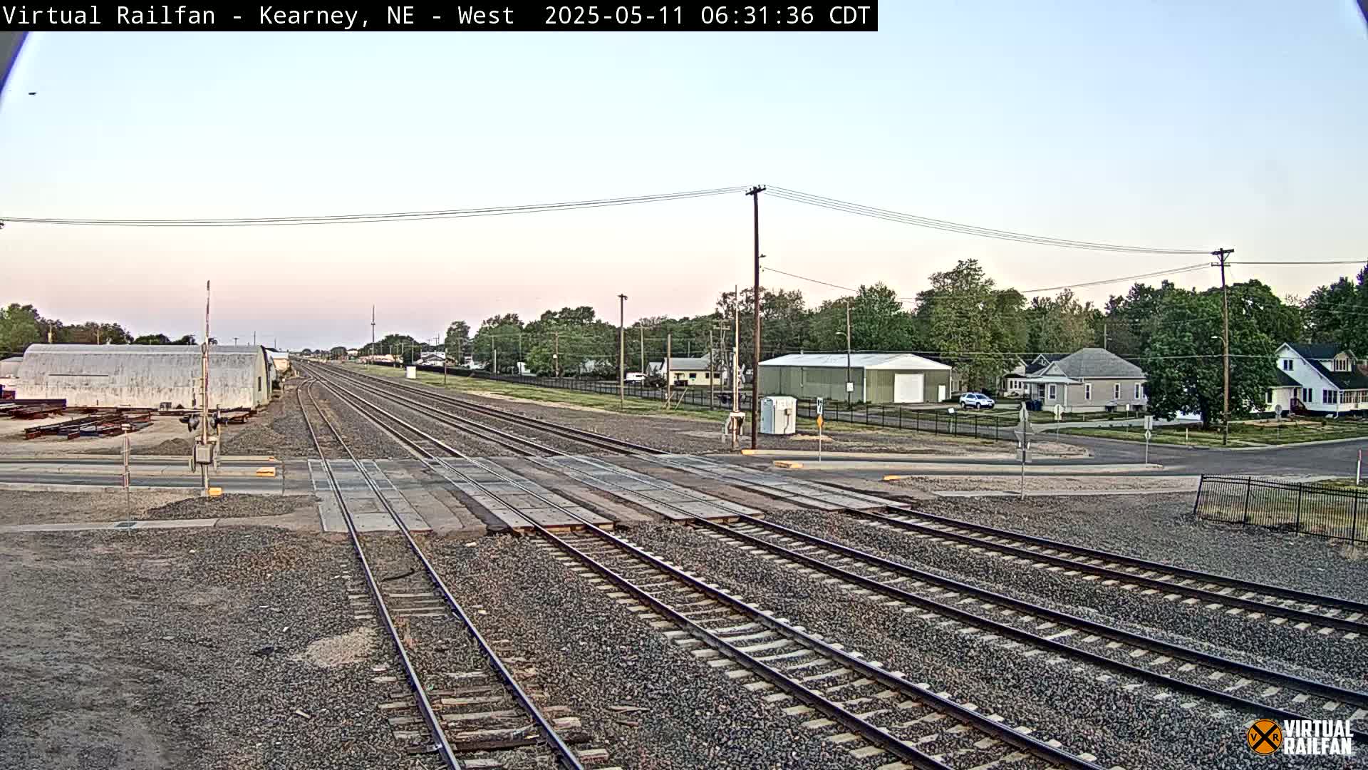 A railroad crossing in a small town during the early morning, under a clear, light sky.