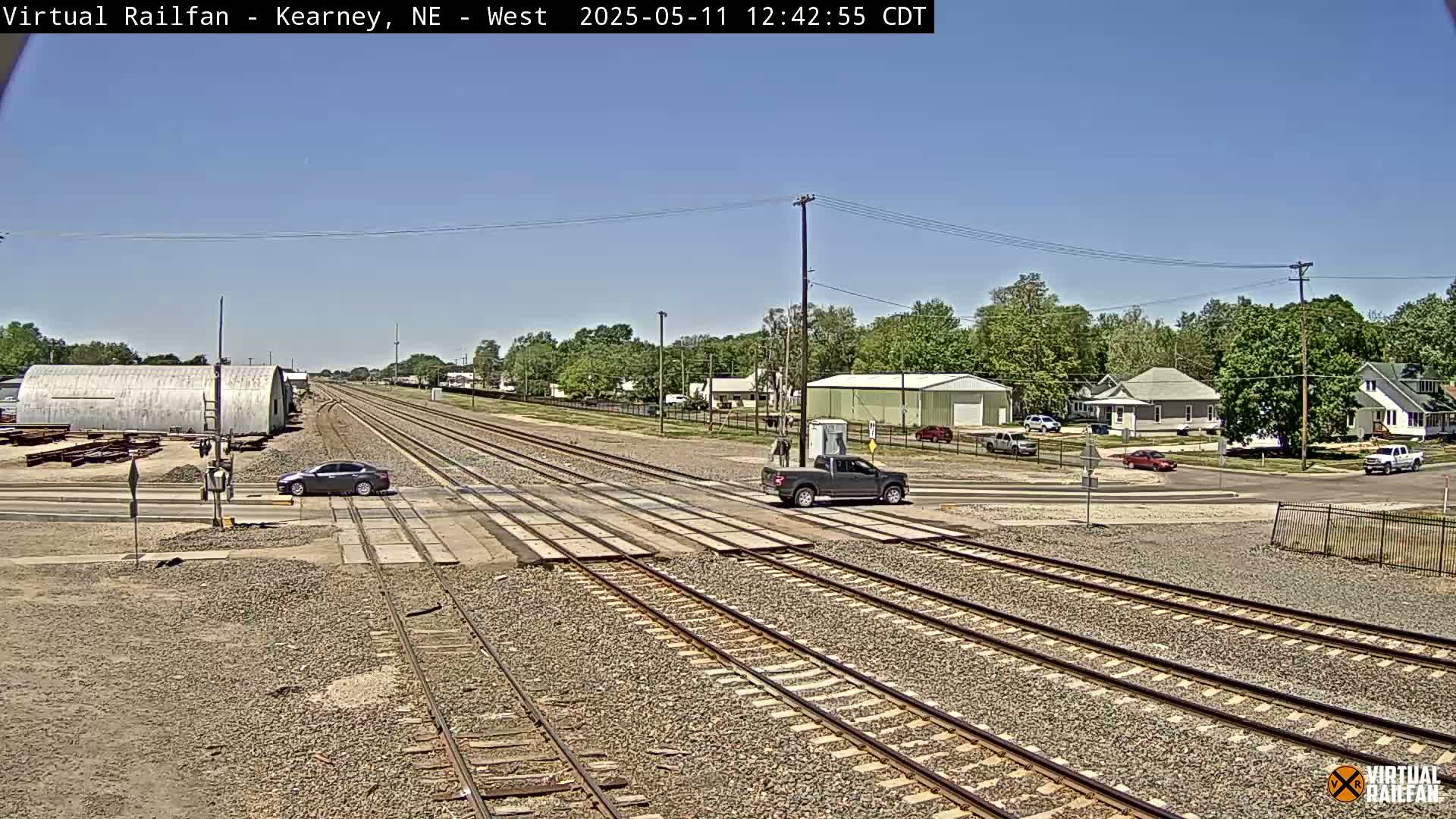 A sunny day reveals railroad tracks intersecting a road, with several vehicles at the crossing and a residential area visible in the background.