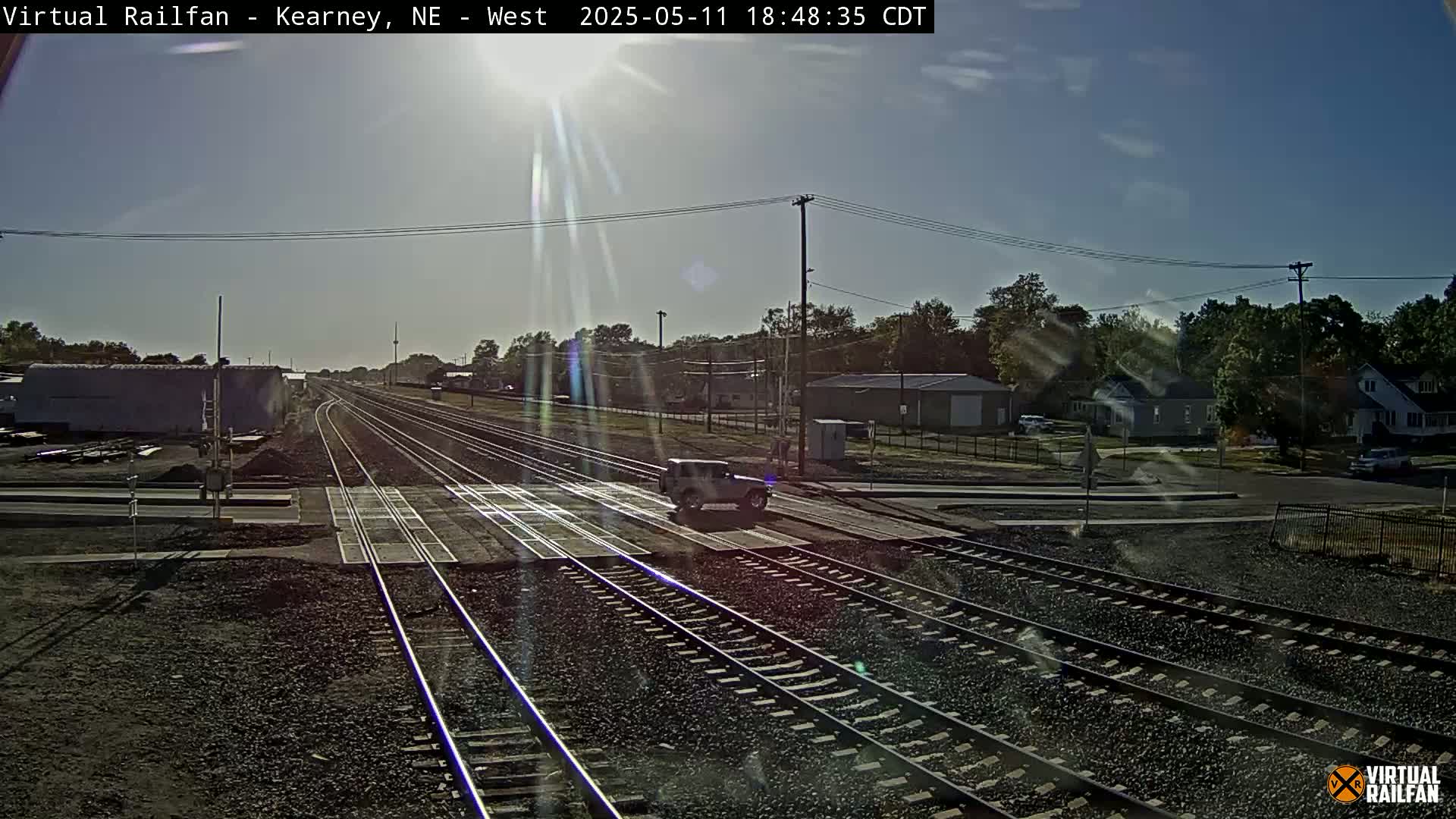 A vehicle is stopped at a railroad crossing under a sunny sky, with trees and buildings visible in the background.
