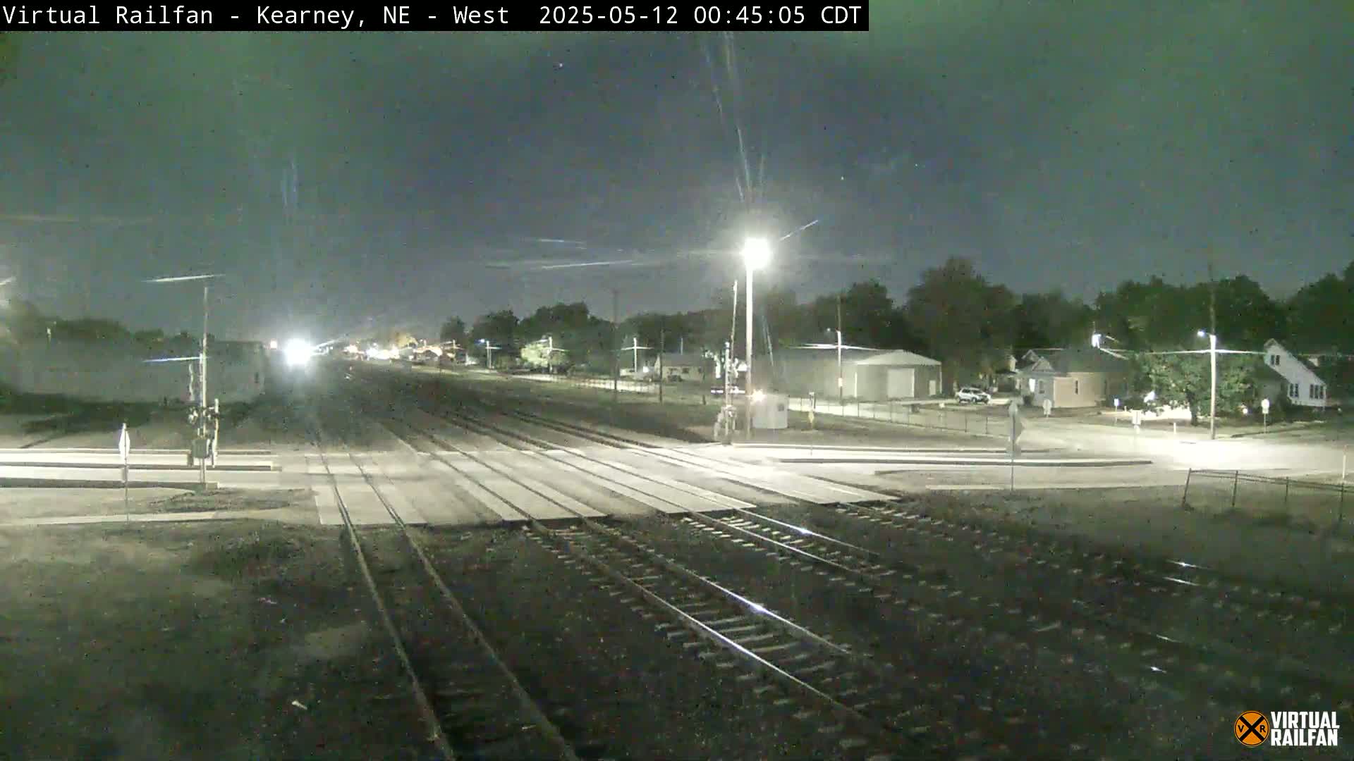 A nighttime view of railroad tracks intersecting with a road, surrounded by residential buildings and trees under a dark sky.
