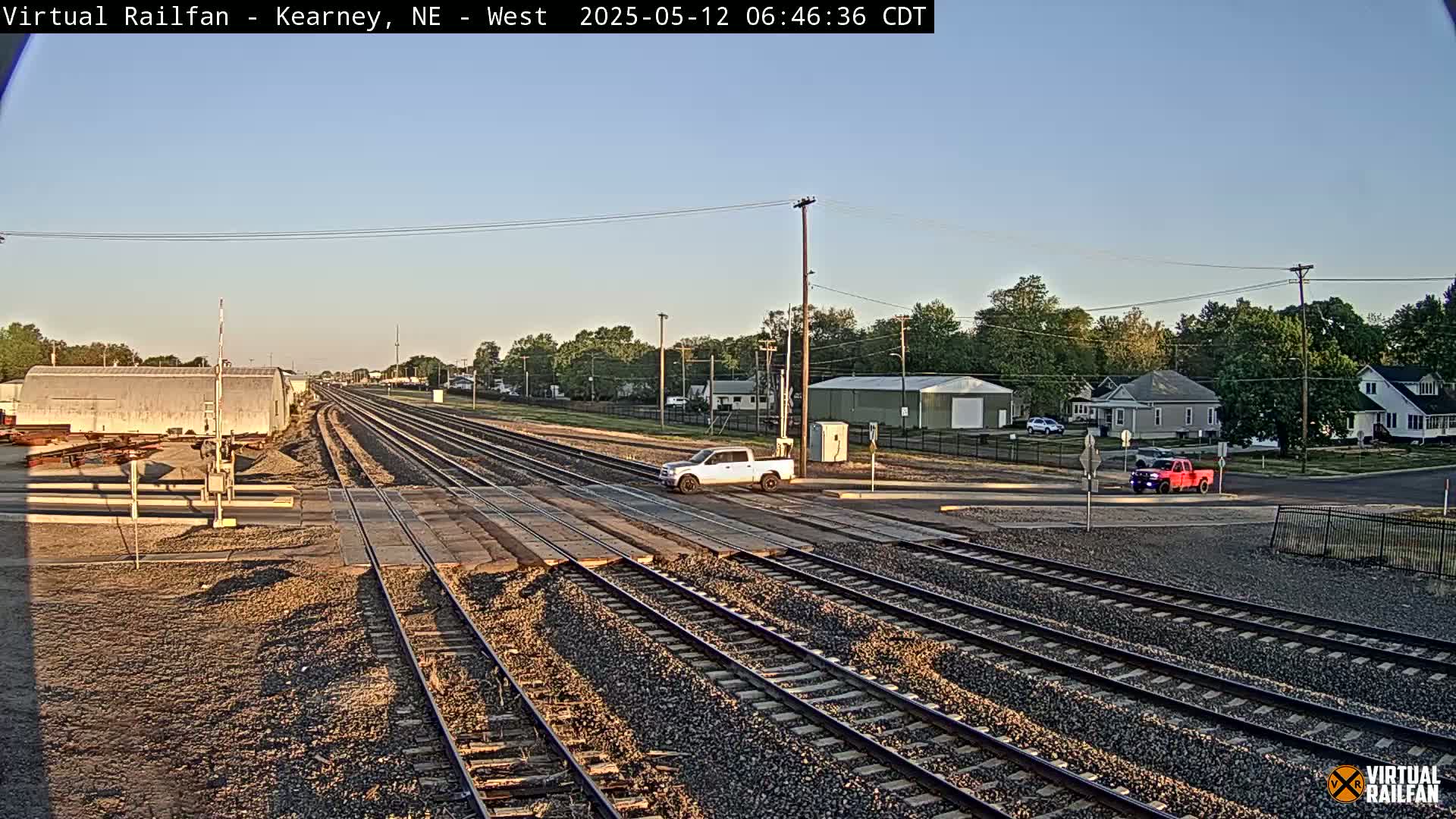 Under a clear sky, a railroad crossing shows multiple train tracks intersecting with a road where a light-colored pickup truck and a red pickup truck are stopped.