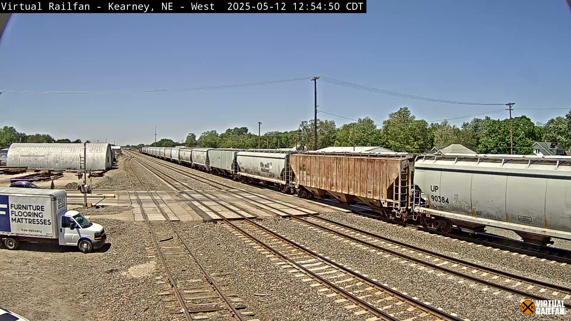 Under a clear blue sky, a long freight train passes through a railroad crossing near a delivery van and industrial buildings.