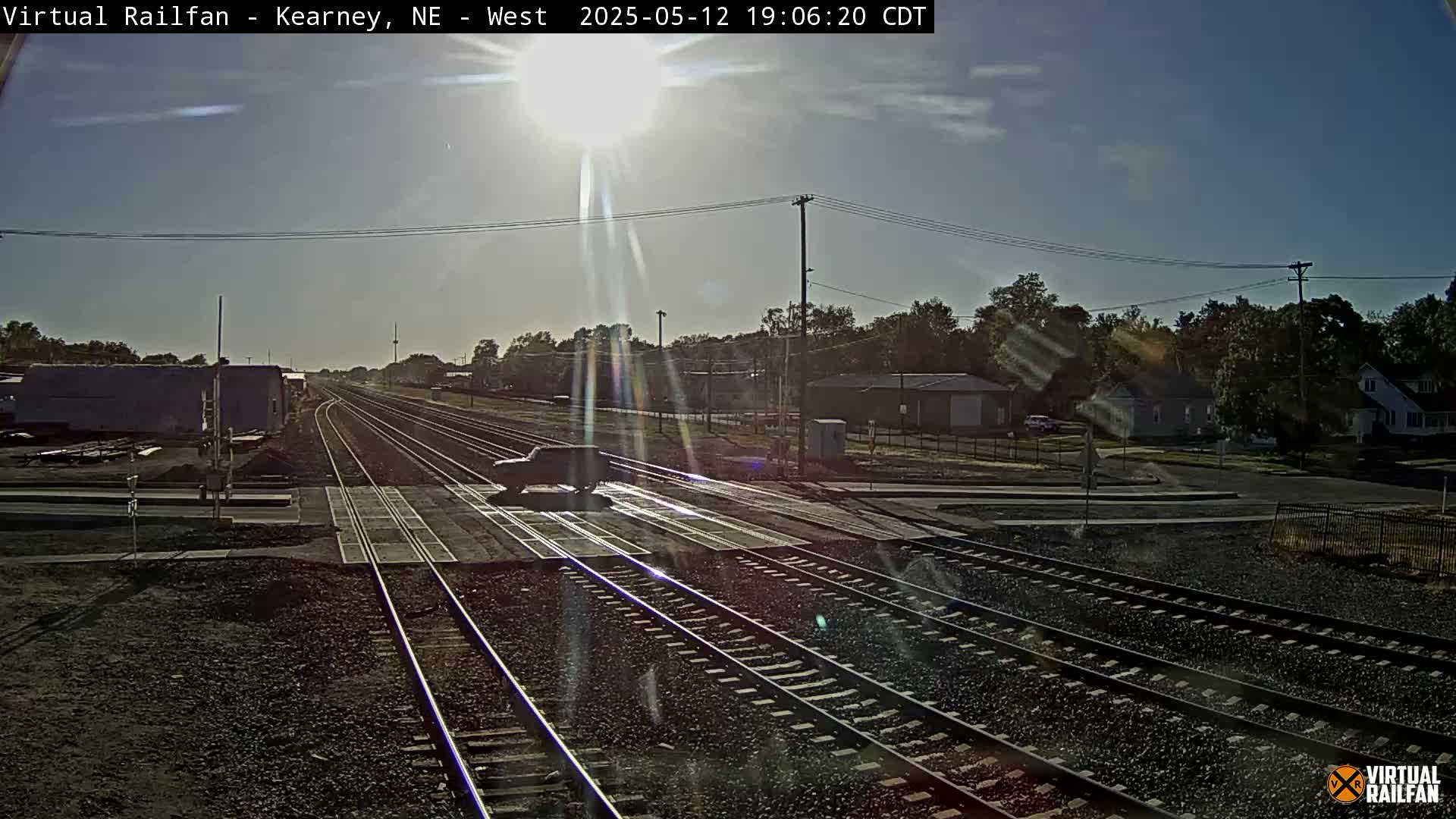 A dark-colored vehicle sits on a sunny railroad crossing near a residential area with several parallel train tracks.