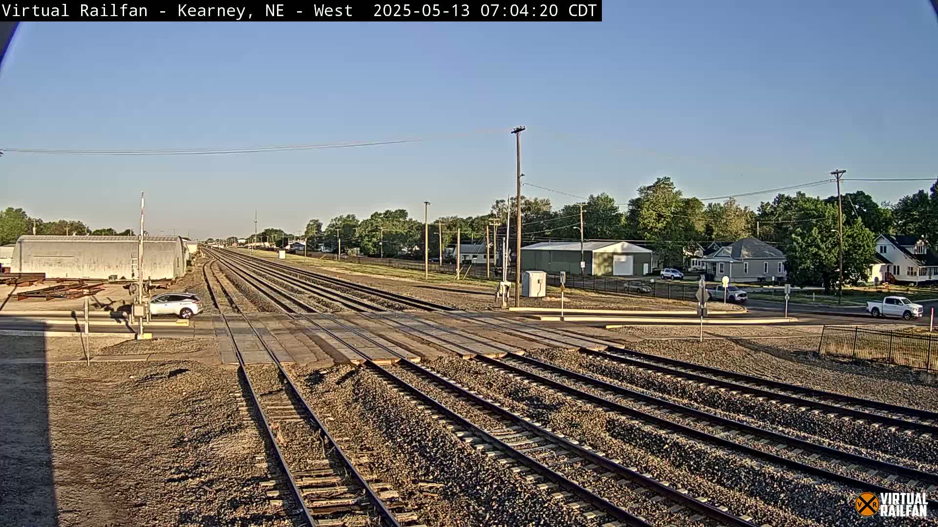 Under a clear sky, multiple train tracks intersect a road near a residential area with a few cars visible.