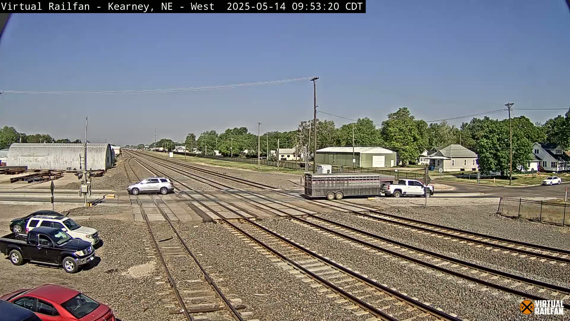 A sunny day reveals multiple vehicles at a railroad crossing near a residential area with trees and buildings in the background.