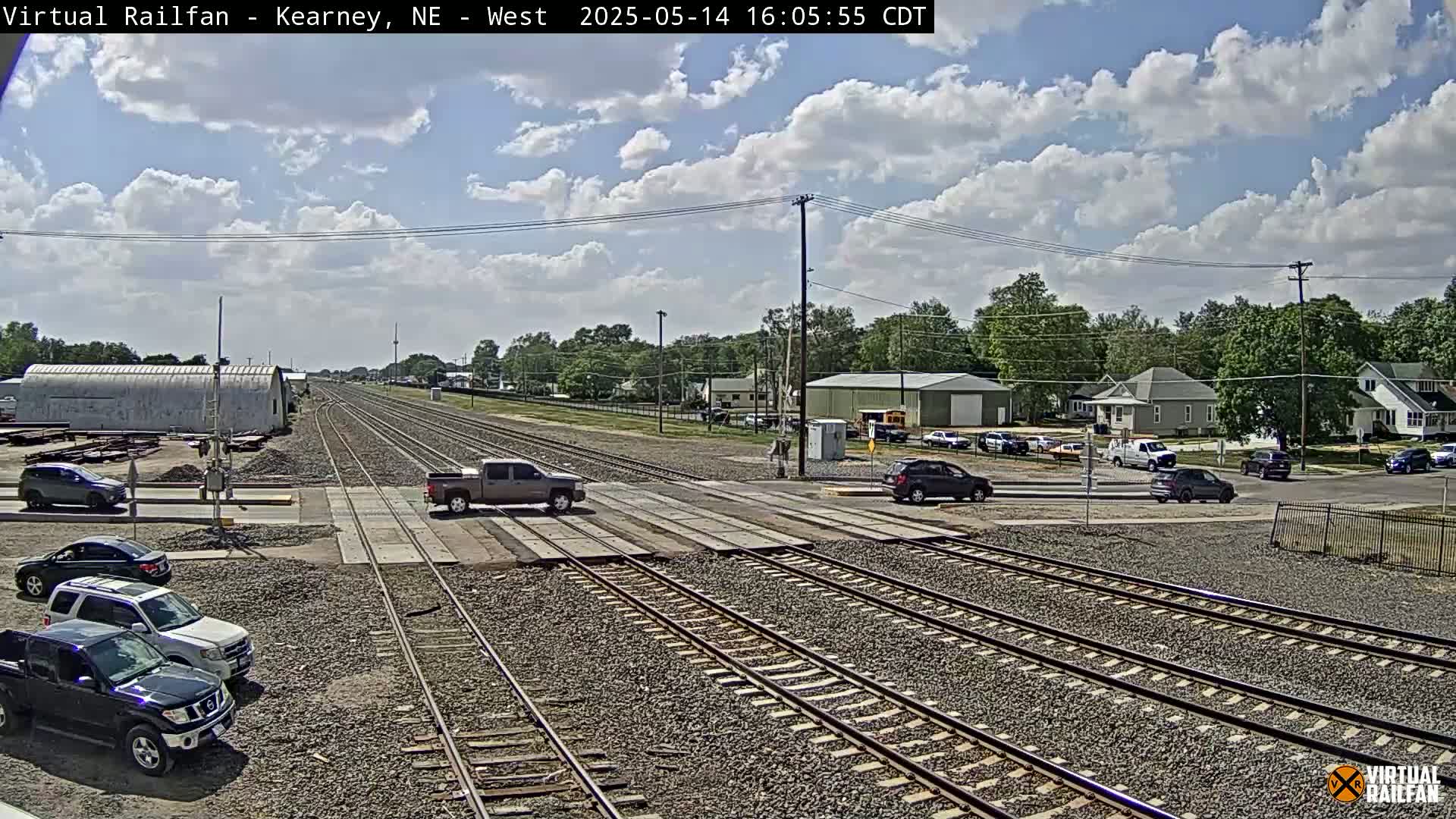 A mostly sunny day with some clouds shows a railroad crossing with several vehicles stopped at the crossing and several more parked nearby, with a small town visible in the background.