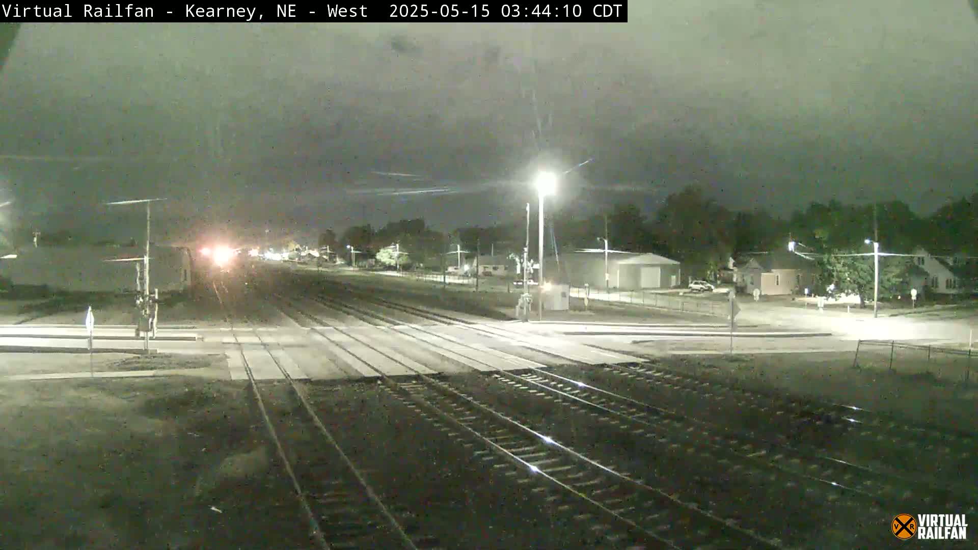 A nighttime view of a railroad crossing with a train in the distance under a cloudy sky.
