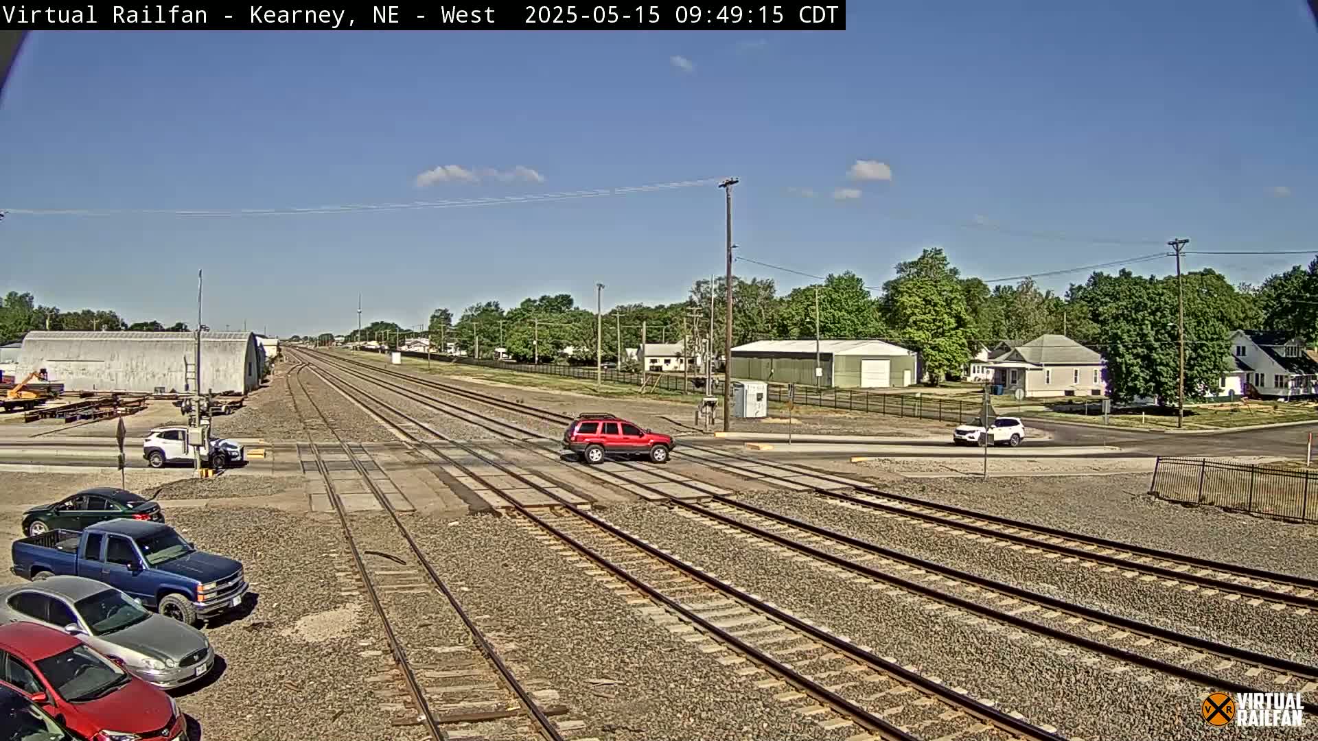 A red SUV drives across a railroad crossing in a town under a clear, sunny sky, with several other vehicles parked nearby.