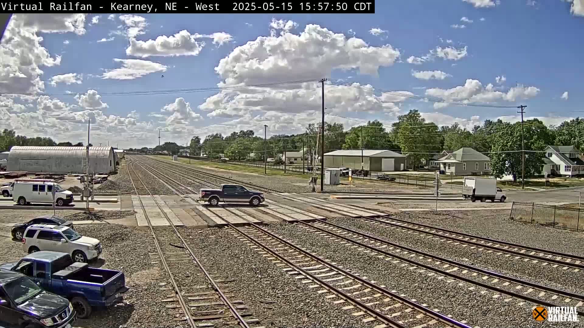 A partly cloudy day shows a railroad crossing with several vehicles parked and driving near train tracks in a residential area.