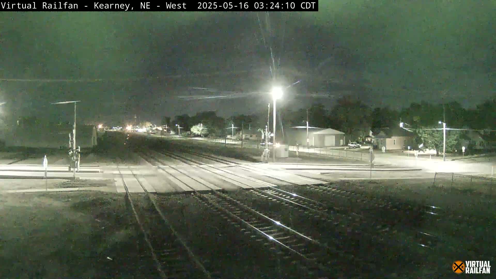 A nighttime view of a railroad crossing with streetlights illuminating train tracks and a nearby residential area under a dark sky.