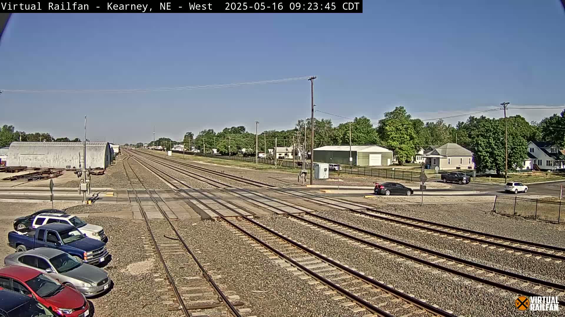 A sunny day reveals railroad tracks intersecting a road in a town, with several cars parked along the side of the road and a few driving on the intersecting road.