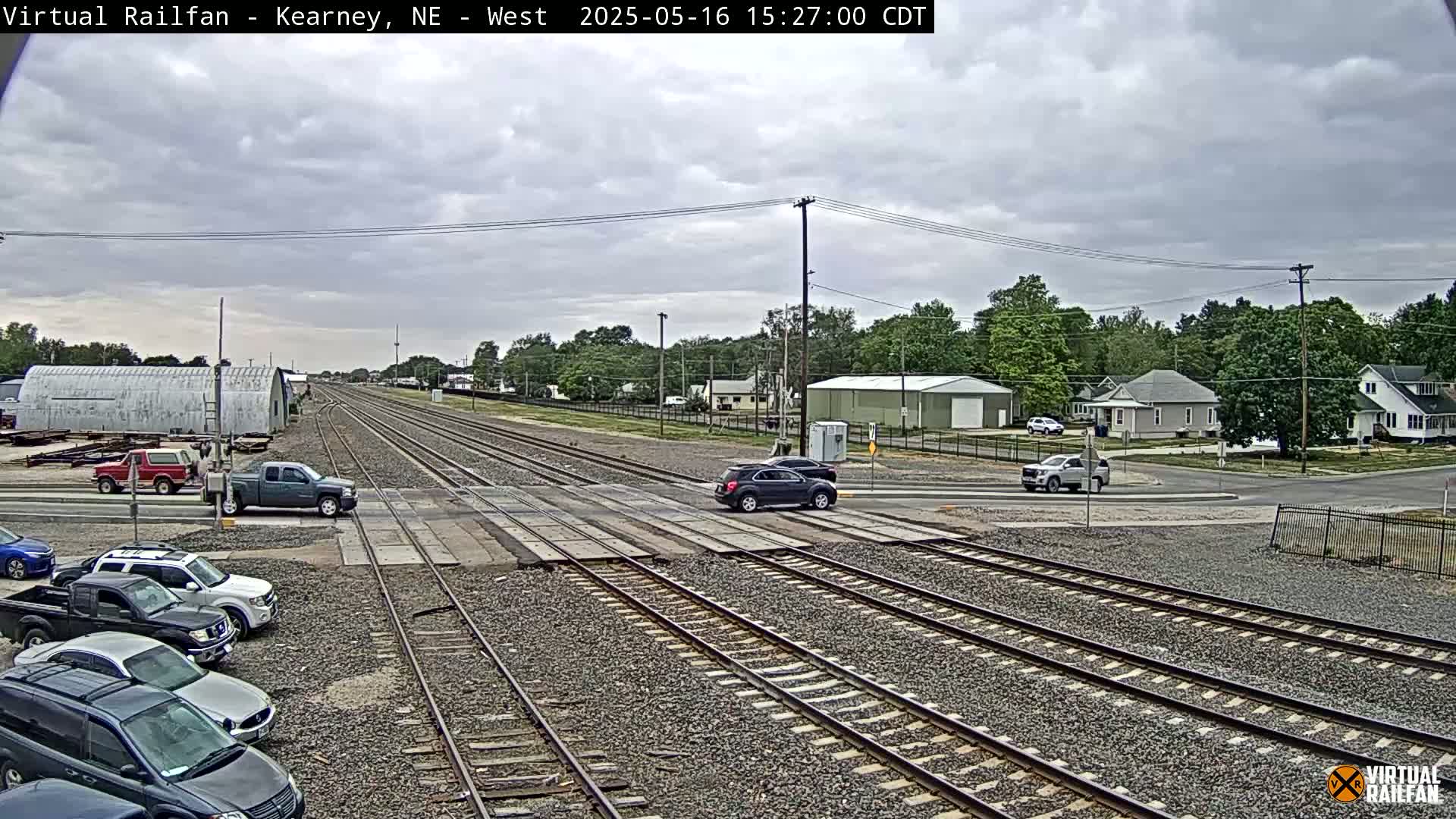 Several vehicles are stopped at a railroad crossing under an overcast sky, with a residential area and industrial buildings visible in the background.
