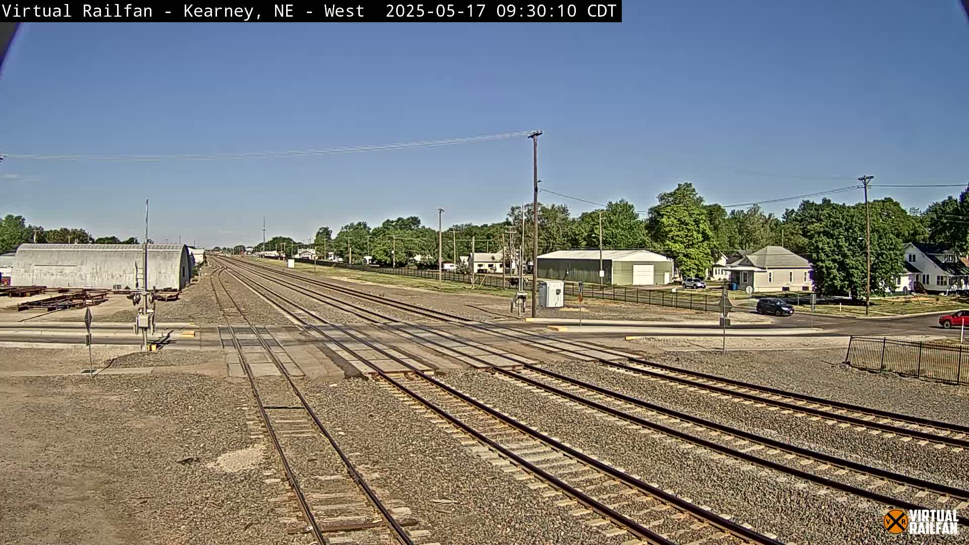 A railroad crossing intersects a road in front of a small town under a clear, sunny sky.