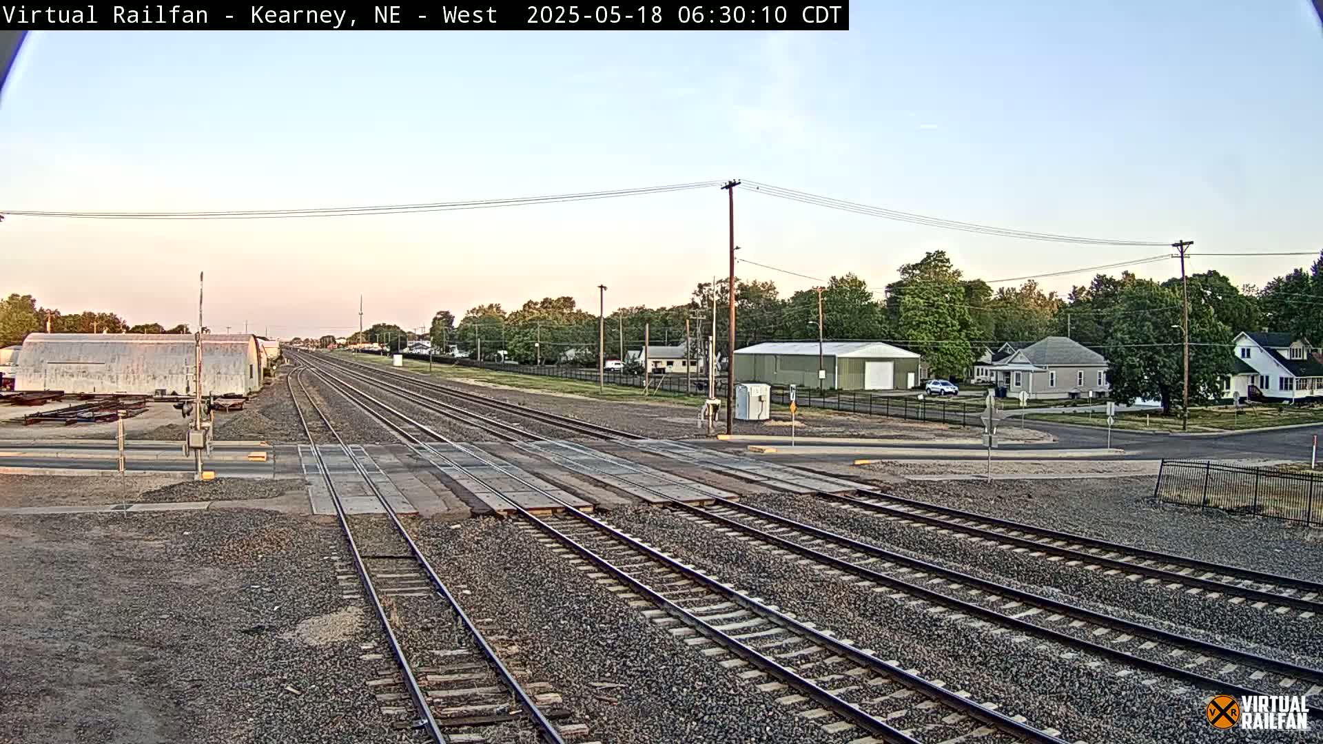 A railroad crossing in a residential area under a clear, twilight sky.