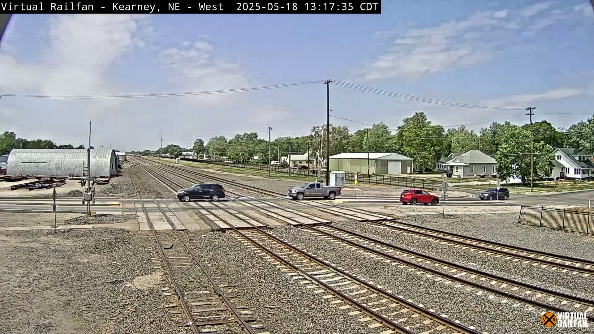 Several vehicles are crossing a railway intersection under a partly cloudy sky in a small town.