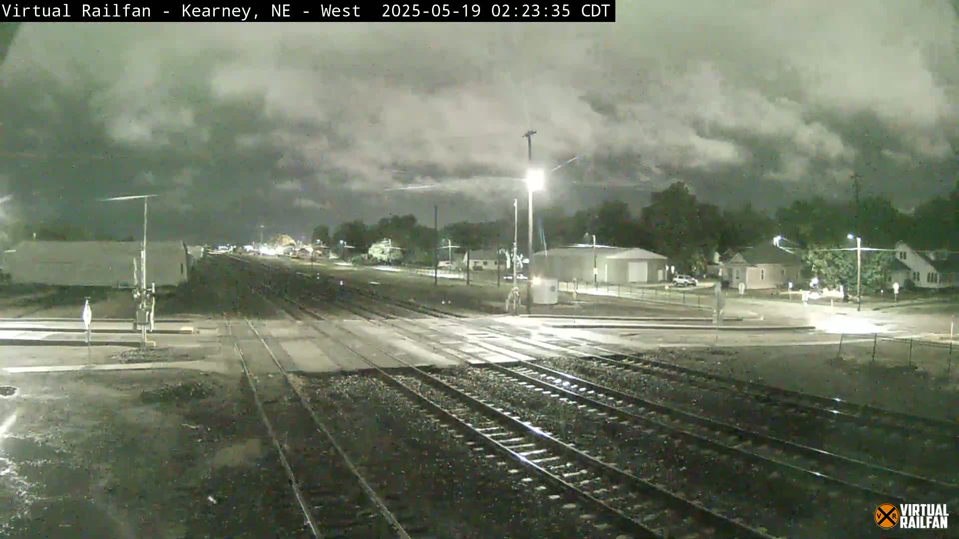 A nighttime view of railroad tracks intersecting a road, under a cloudy sky, with residential buildings and streetlights visible in the background.