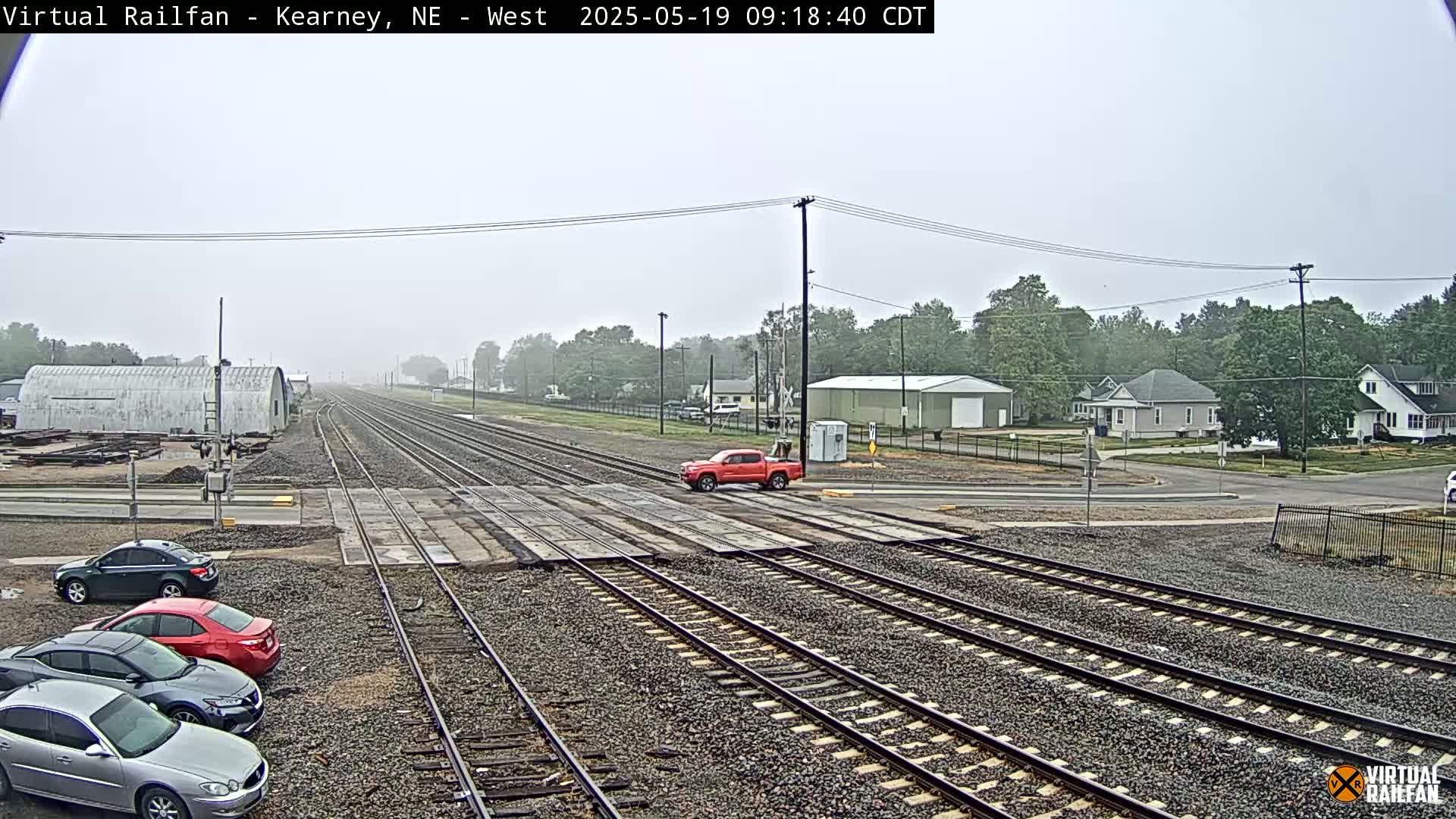 A foggy scene shows multiple sets of train tracks intersecting a road with several cars parked along the side and a red pickup truck stopped at the tracks.