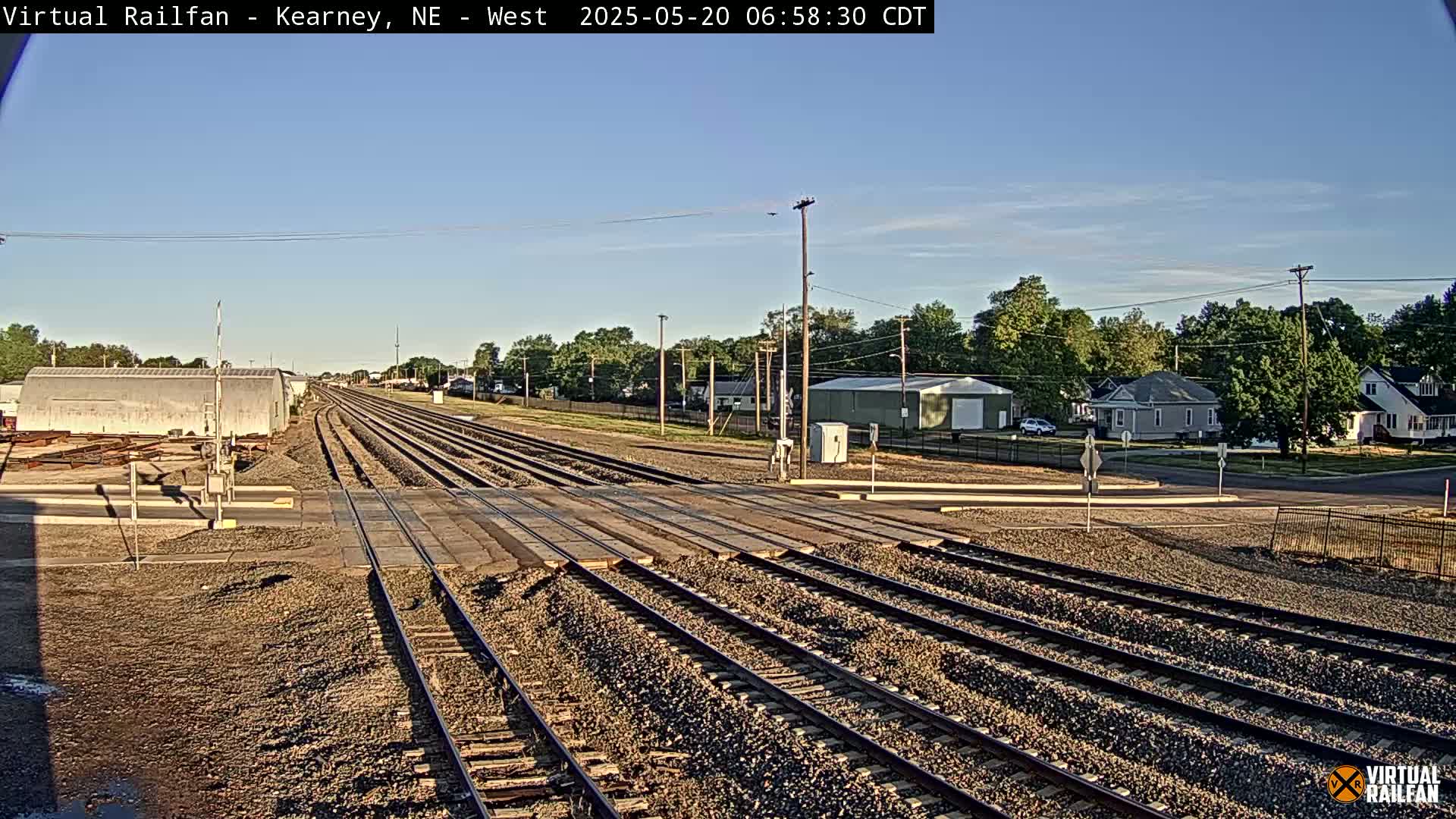 A railroad crossing with multiple train tracks is seen on a sunny day, with buildings and trees visible in the background.