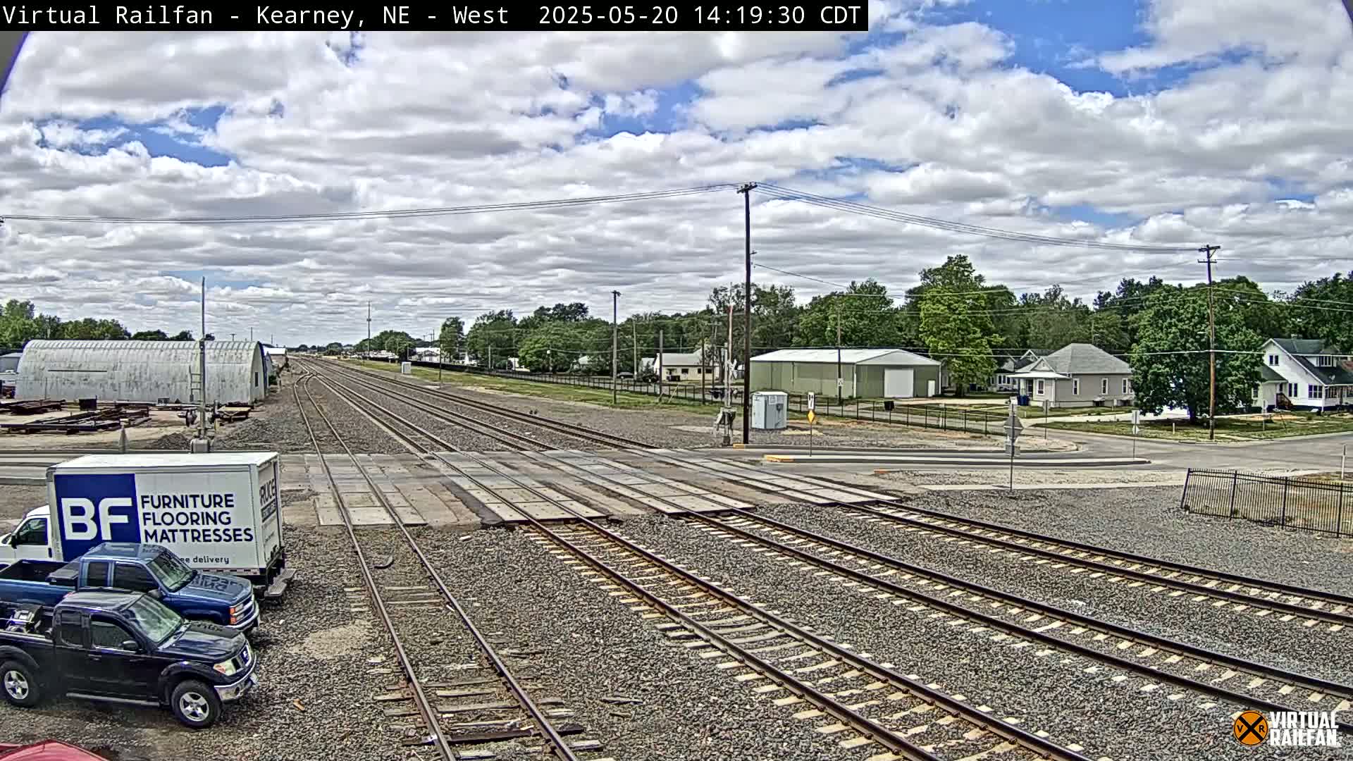 Under a partly cloudy sky, railroad tracks cross a road in front of several buildings and parked trucks.