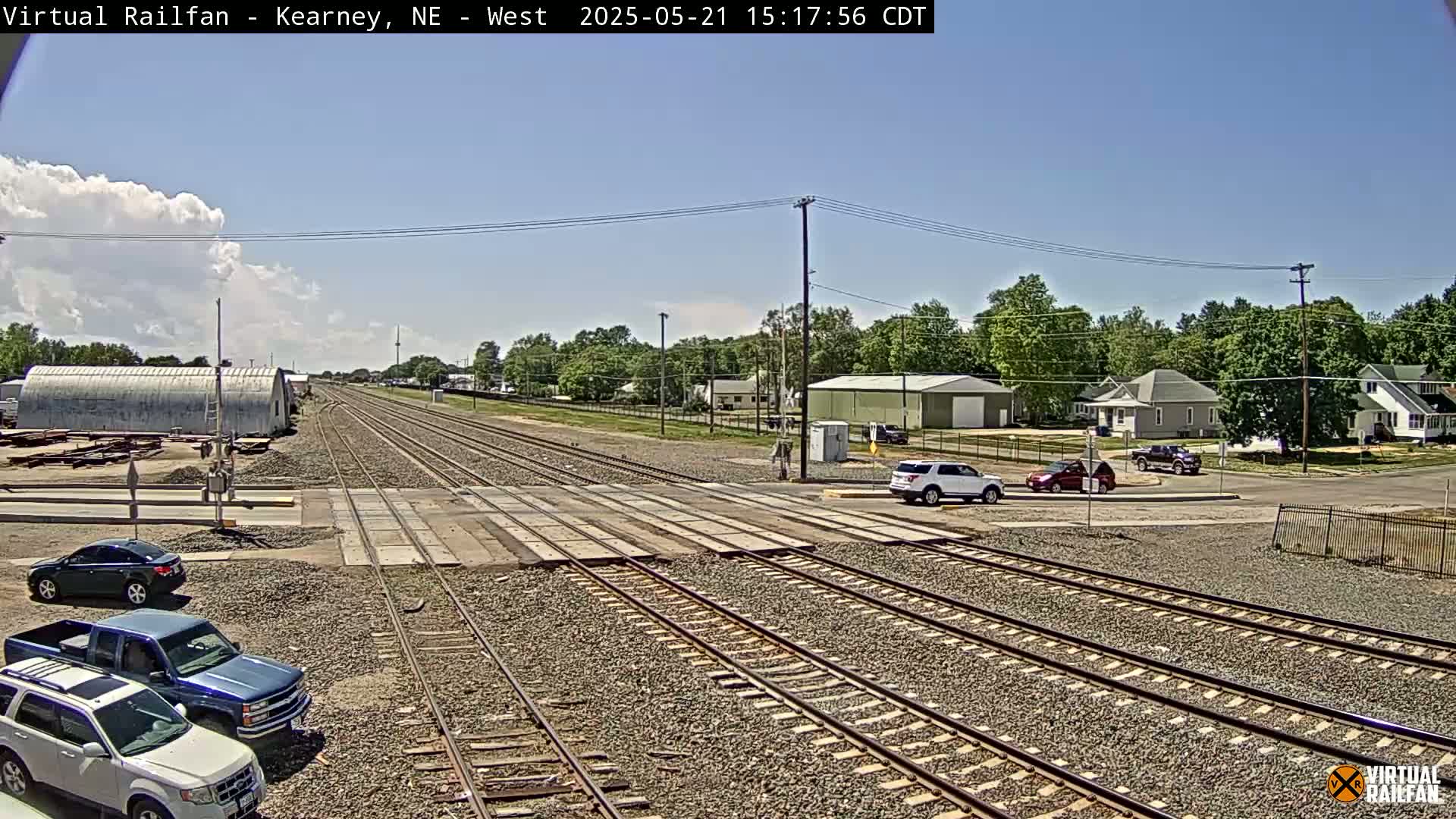 A railroad crossing in a town under partly cloudy skies shows several vehicles stopped near the tracks, with residential buildings and trees visible in the background.