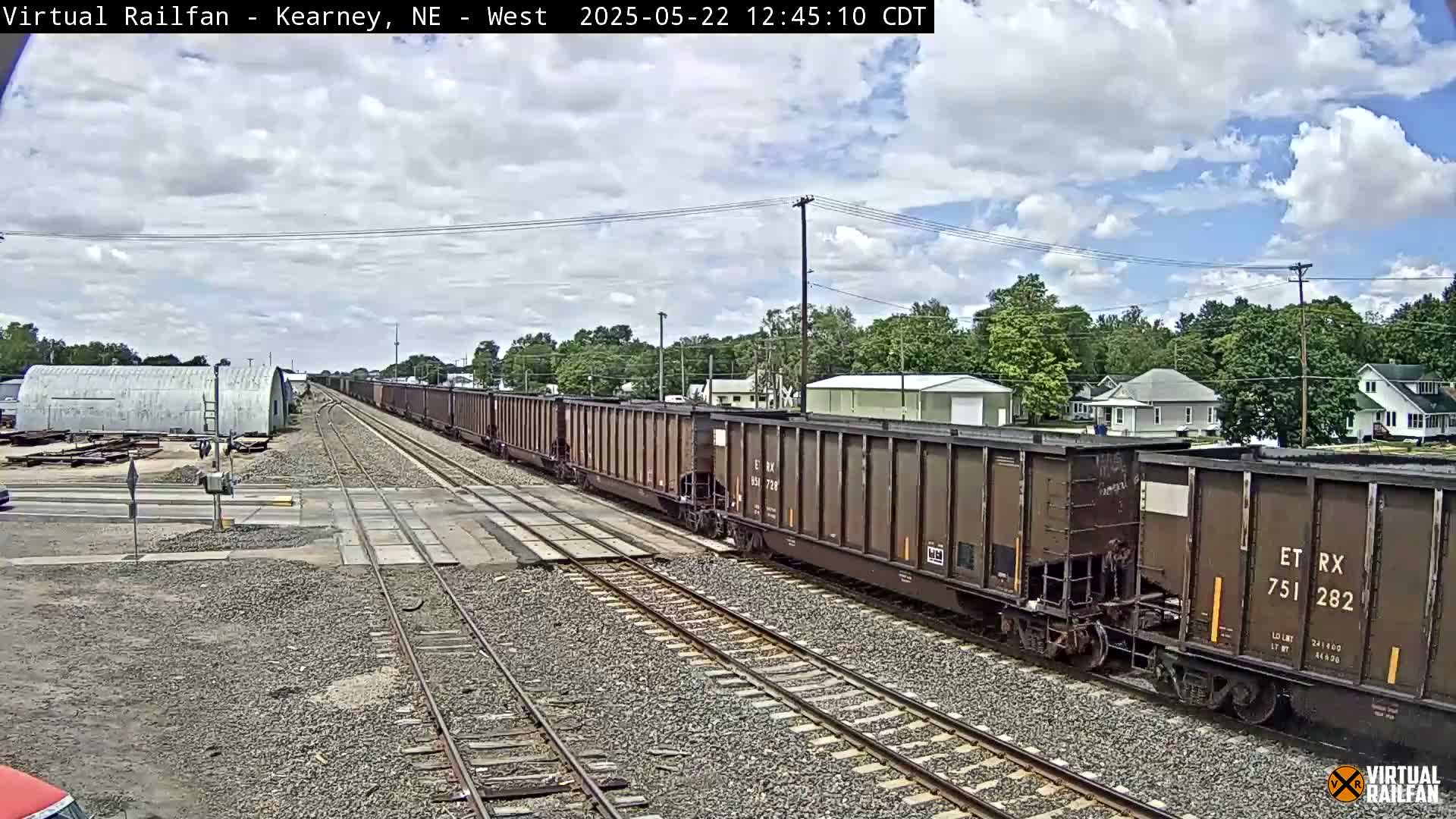 A long freight train passes through a railroad crossing under a partly cloudy sky.
