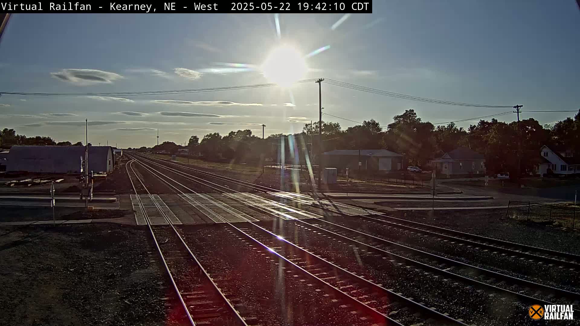 Multiple train tracks intersect a road near houses under a bright, sunny sky with a few clouds.