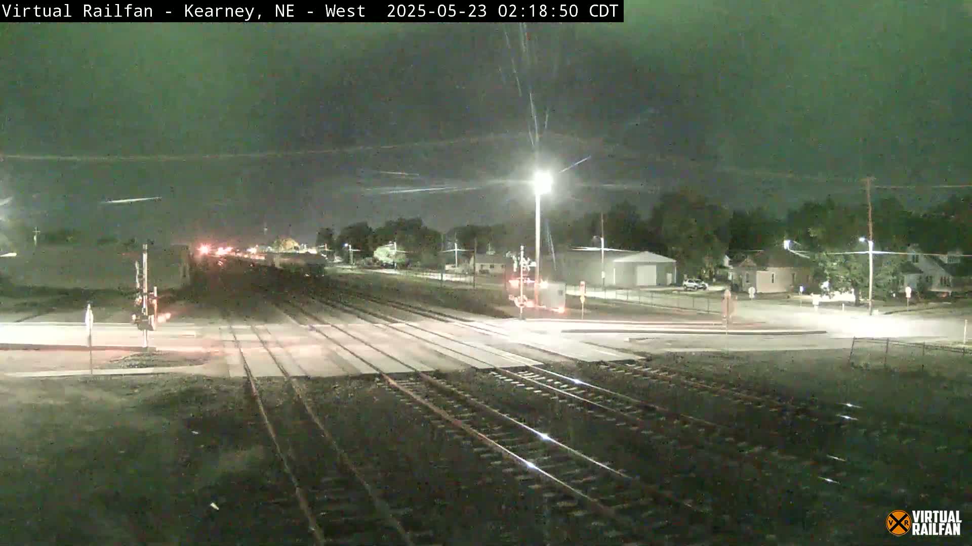 A nighttime view of a railroad crossing with train tracks intersecting a road, under a dark sky with some light cloud cover.