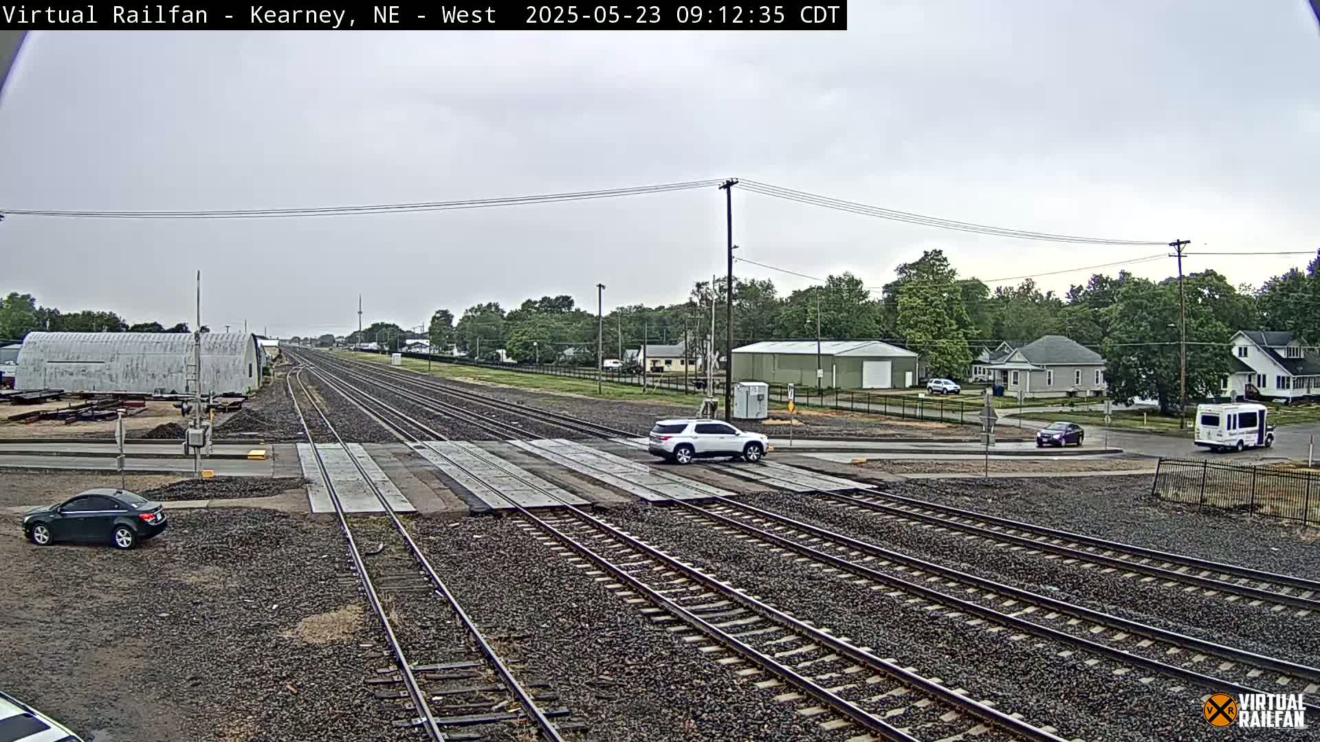 A railroad crossing with several sets of tracks, a few cars at the crossing and nearby roads, and a residential area in the background under an overcast sky.