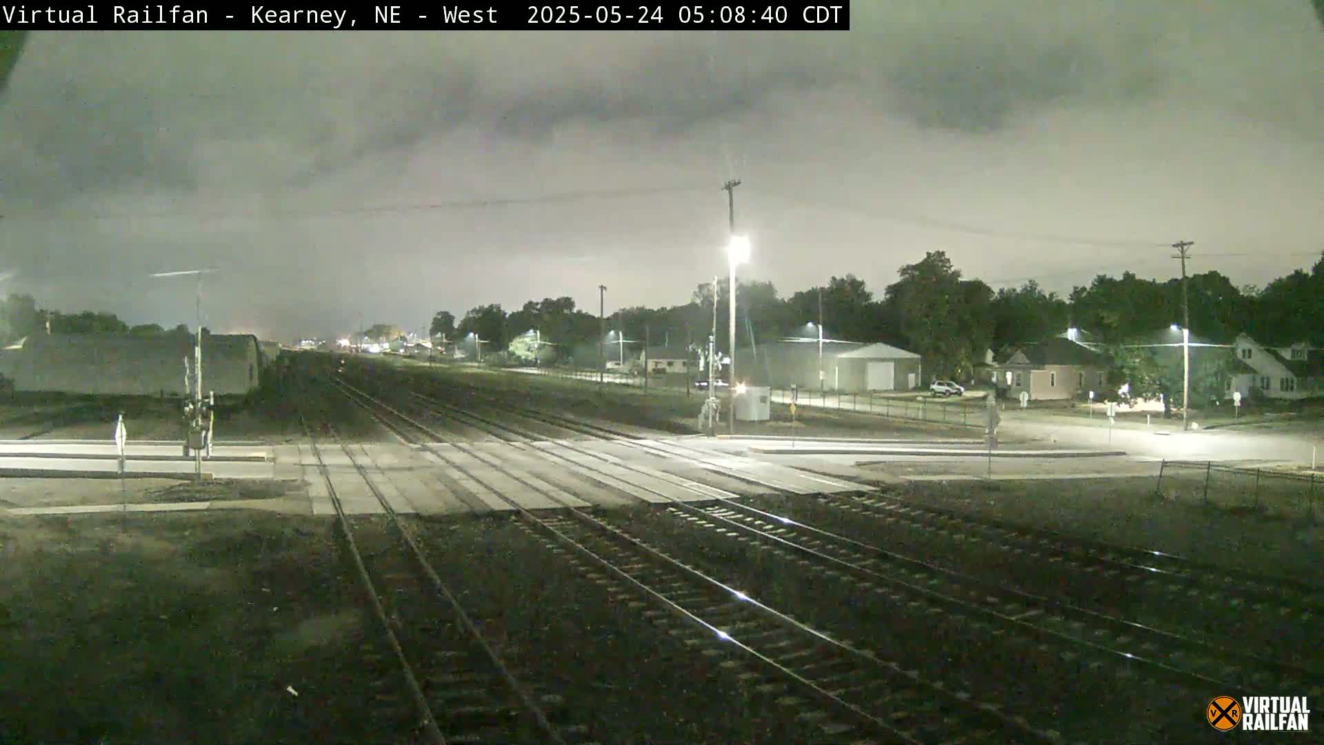 A nighttime view of a railroad crossing in a residential area under a cloudy sky.