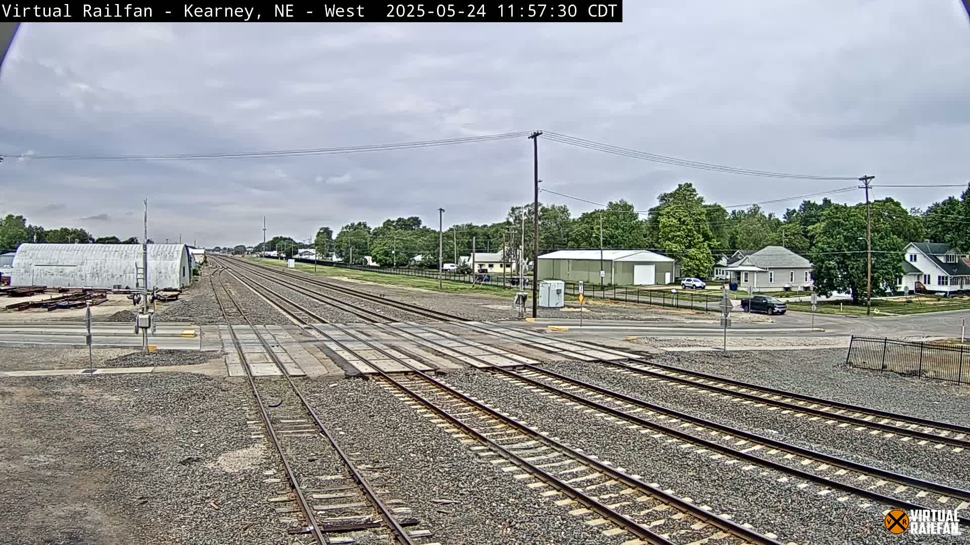 A railroad crossing intersects a road in a residential area under a partly cloudy sky.