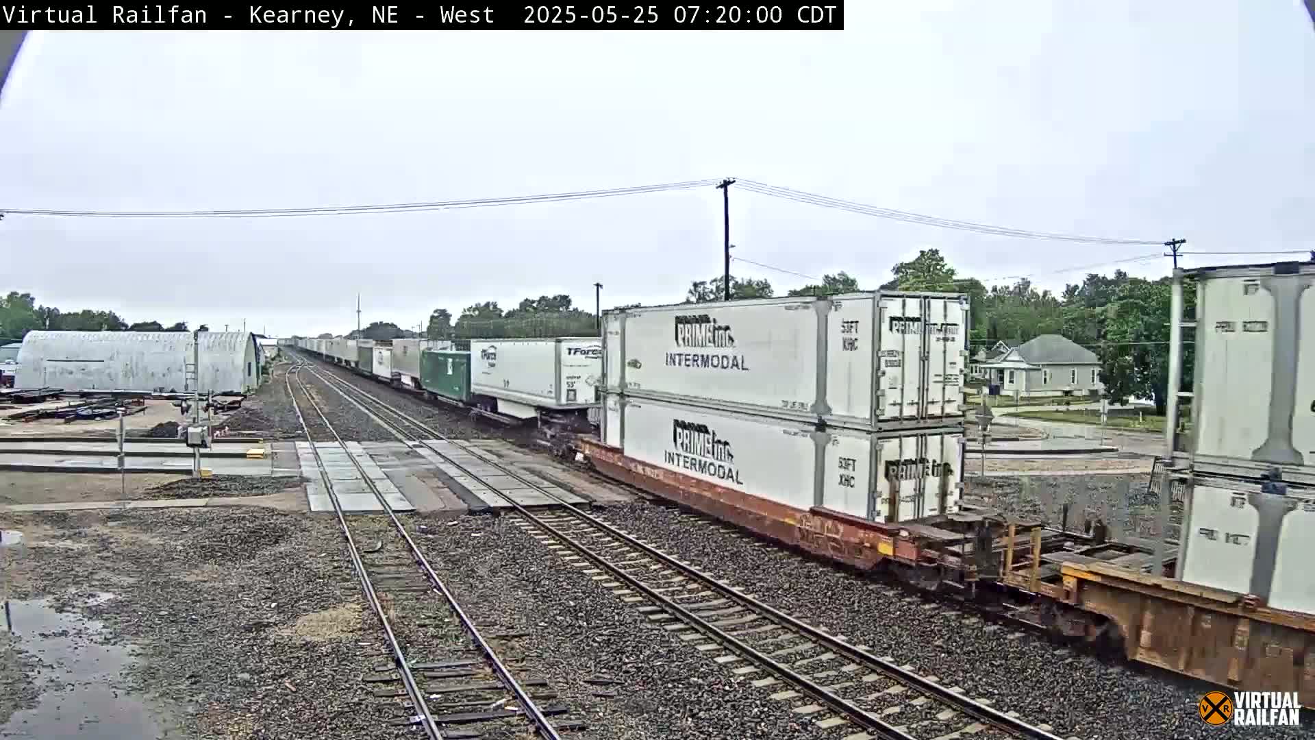 A freight train carrying stacked shipping containers passes through a railroad crossing on an overcast day.