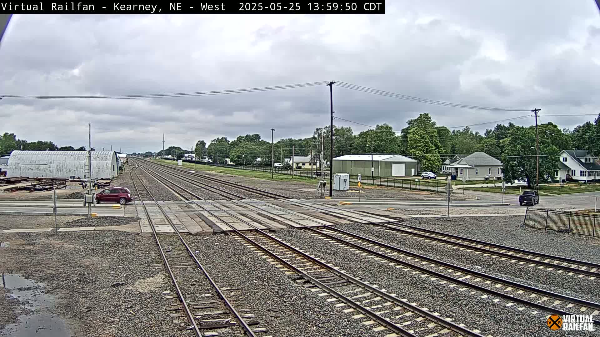 A railroad crossing with several train tracks, a red SUV, and a few buildings under a cloudy sky.