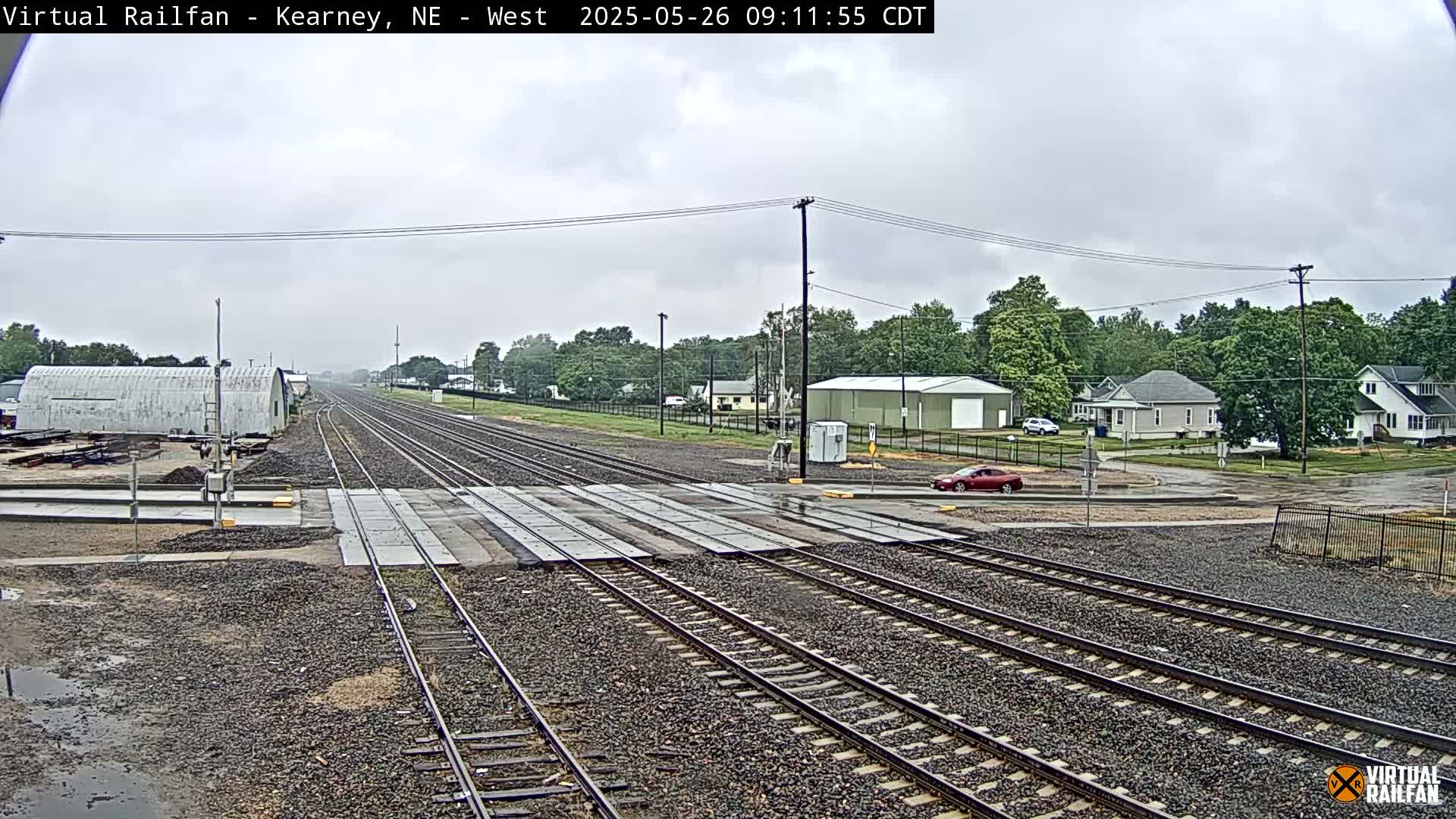 A railroad crossing with several sets of tracks stretches toward a residential area under an overcast sky.