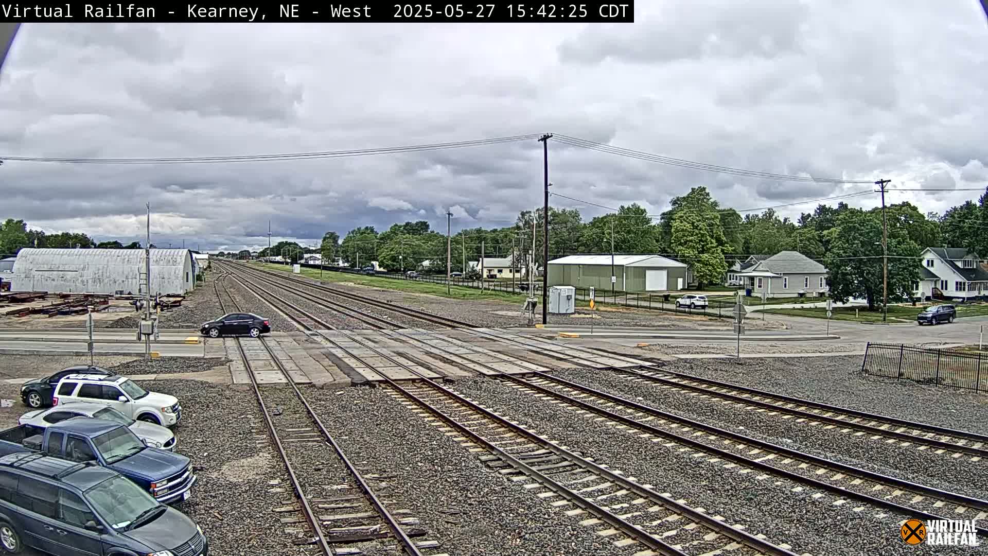 A mostly cloudy day shows several cars parked near railroad tracks that cross a road in front of residential buildings and some industrial structures.