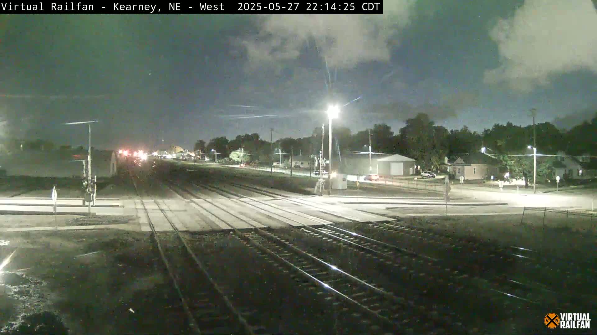 A nighttime view of a railroad crossing, with a street intersecting the tracks under partly cloudy skies.