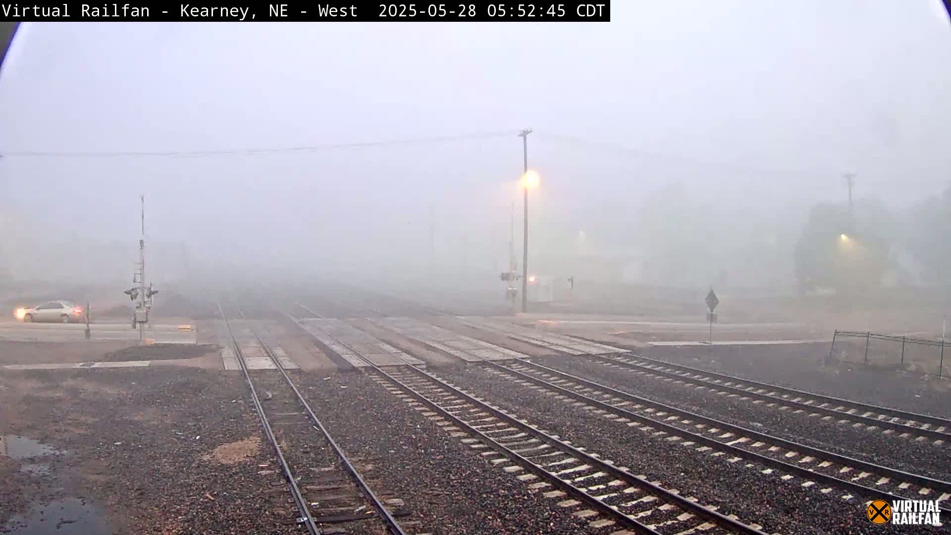 A foggy scene shows several sets of train tracks intersecting with a road, where a vehicle is visible in the distance.