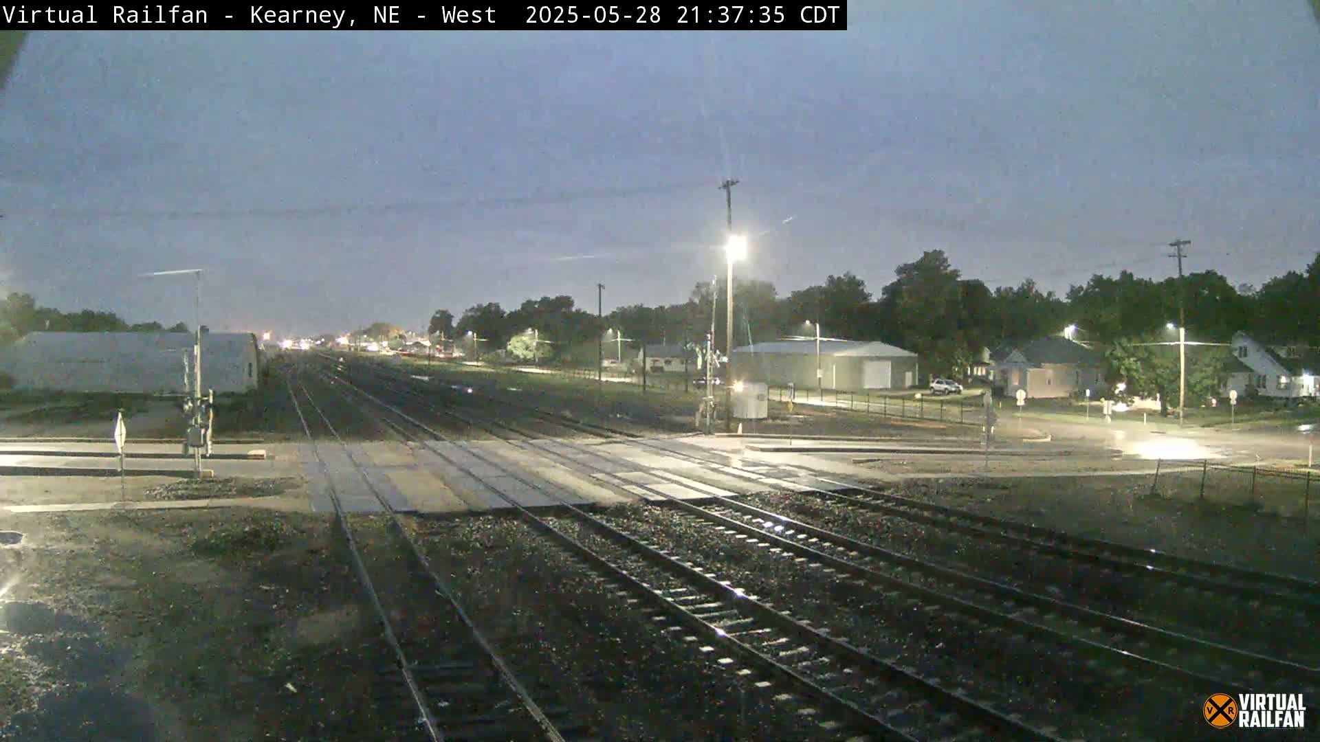 A nighttime view of railroad tracks intersecting with a road, under a dark, possibly overcast sky, with buildings and trees visible in the background.
