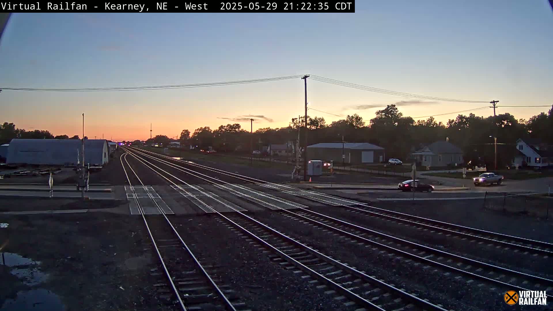 Multiple train tracks converge at a road crossing during a twilight sunset, with a few cars visible at the intersection and some buildings in the background.