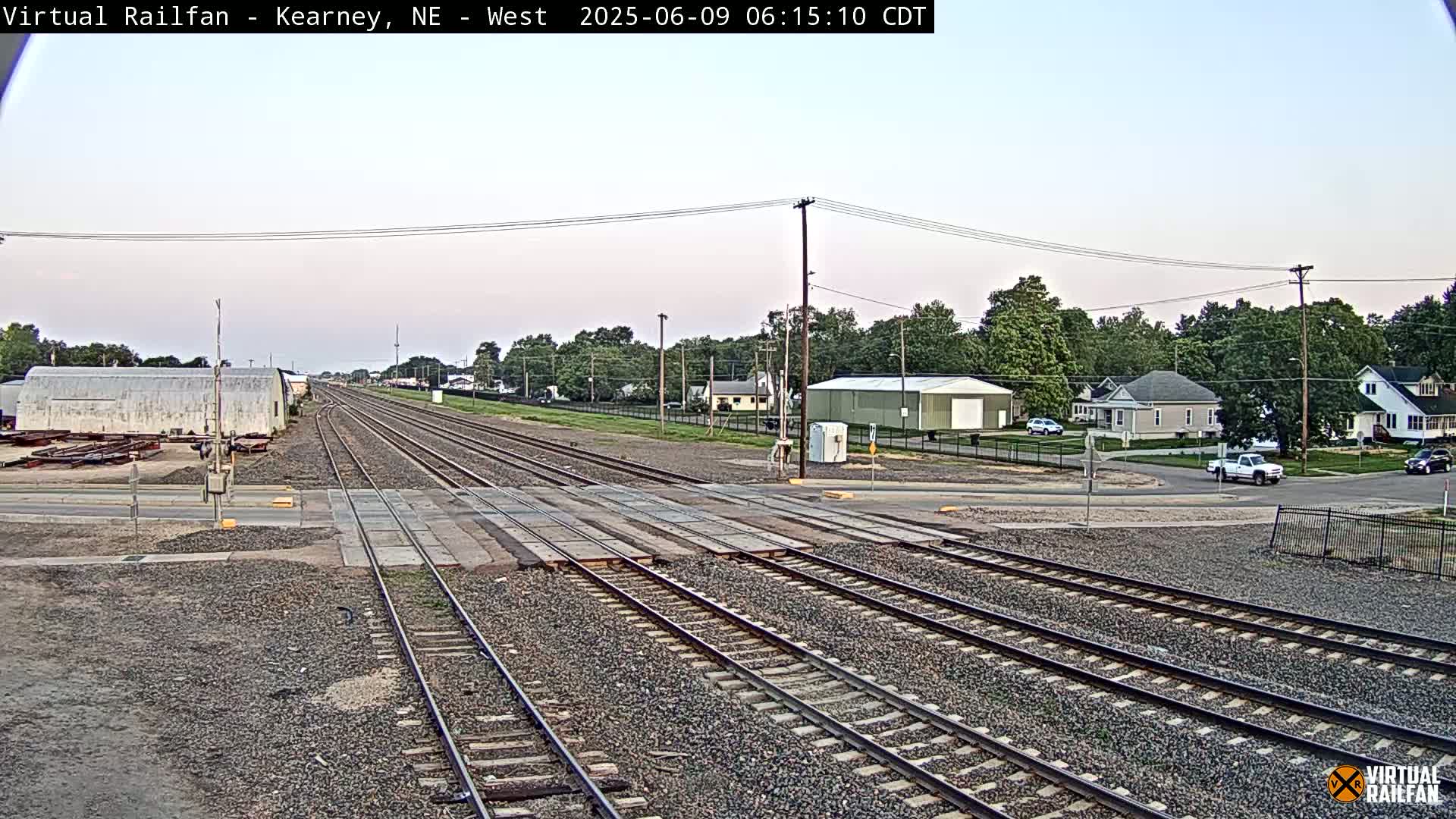 A railroad crossing in a residential area on a partly cloudy day shows multiple train tracks intersecting a road with several houses and vehicles visible in the background.