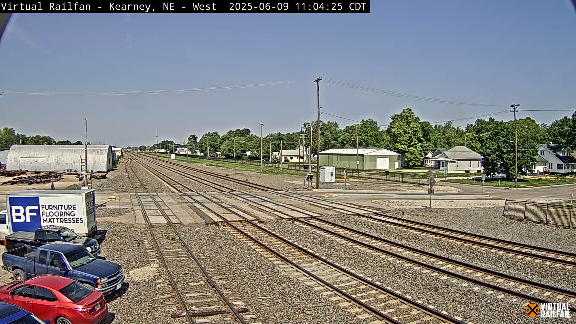 Under a clear blue sky, a railroad track stretches towards a small town, with several parked vehicles and buildings visible.