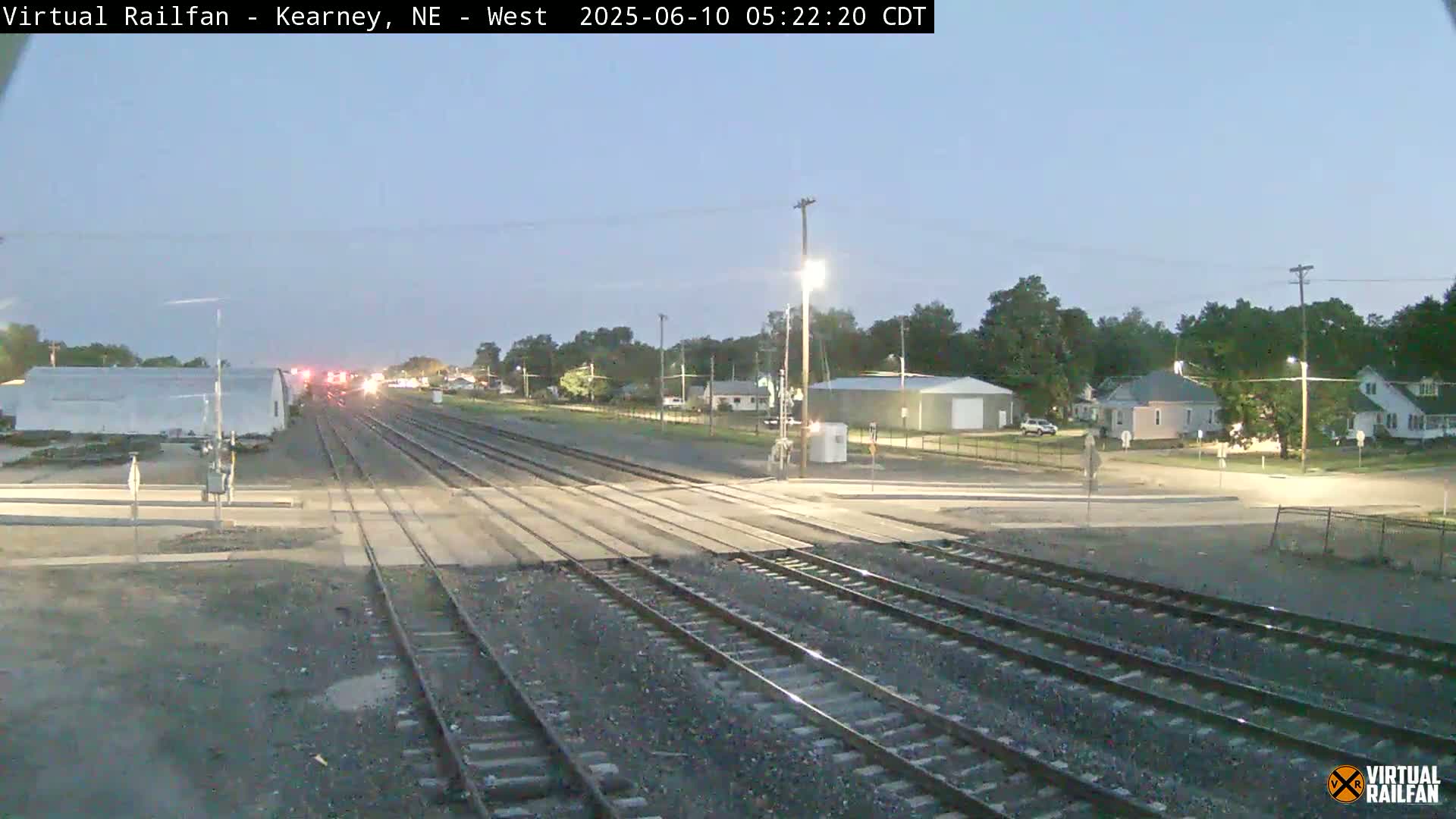 A twilight view of a railway crossing in a small town, with several buildings visible in the background.