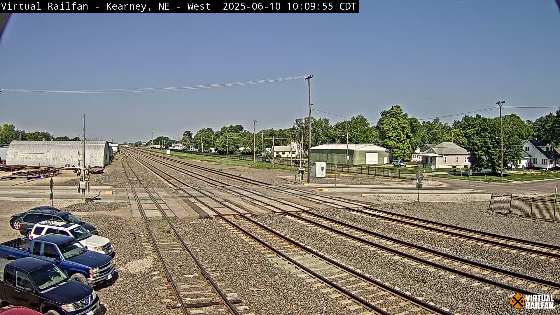 Under a clear blue sky, railroad tracks intersect a road in front of a small town with several parked cars visible near the tracks.